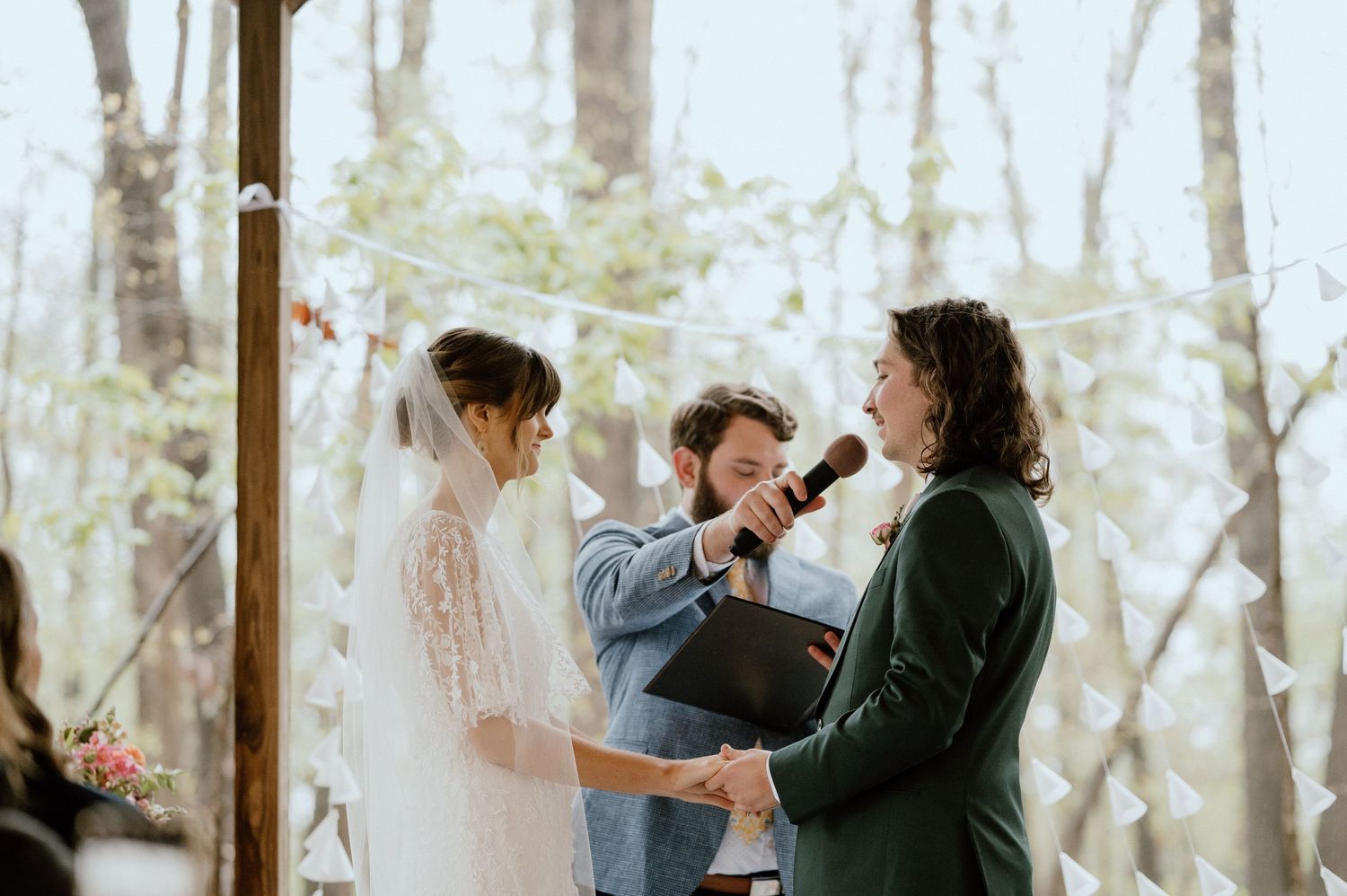 A wedding ceremony takes place in a wooded setting with a couple and officiant sharing an emotional moment.