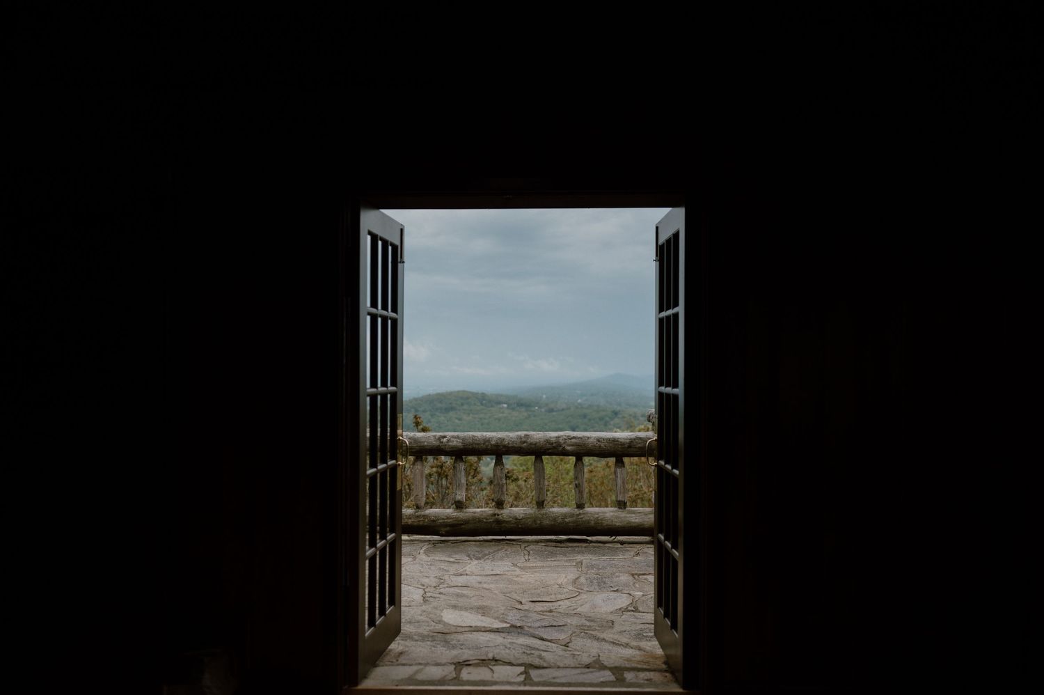 Open rustic wooden doors frame a scenic mountain vista against a cloudy sky.