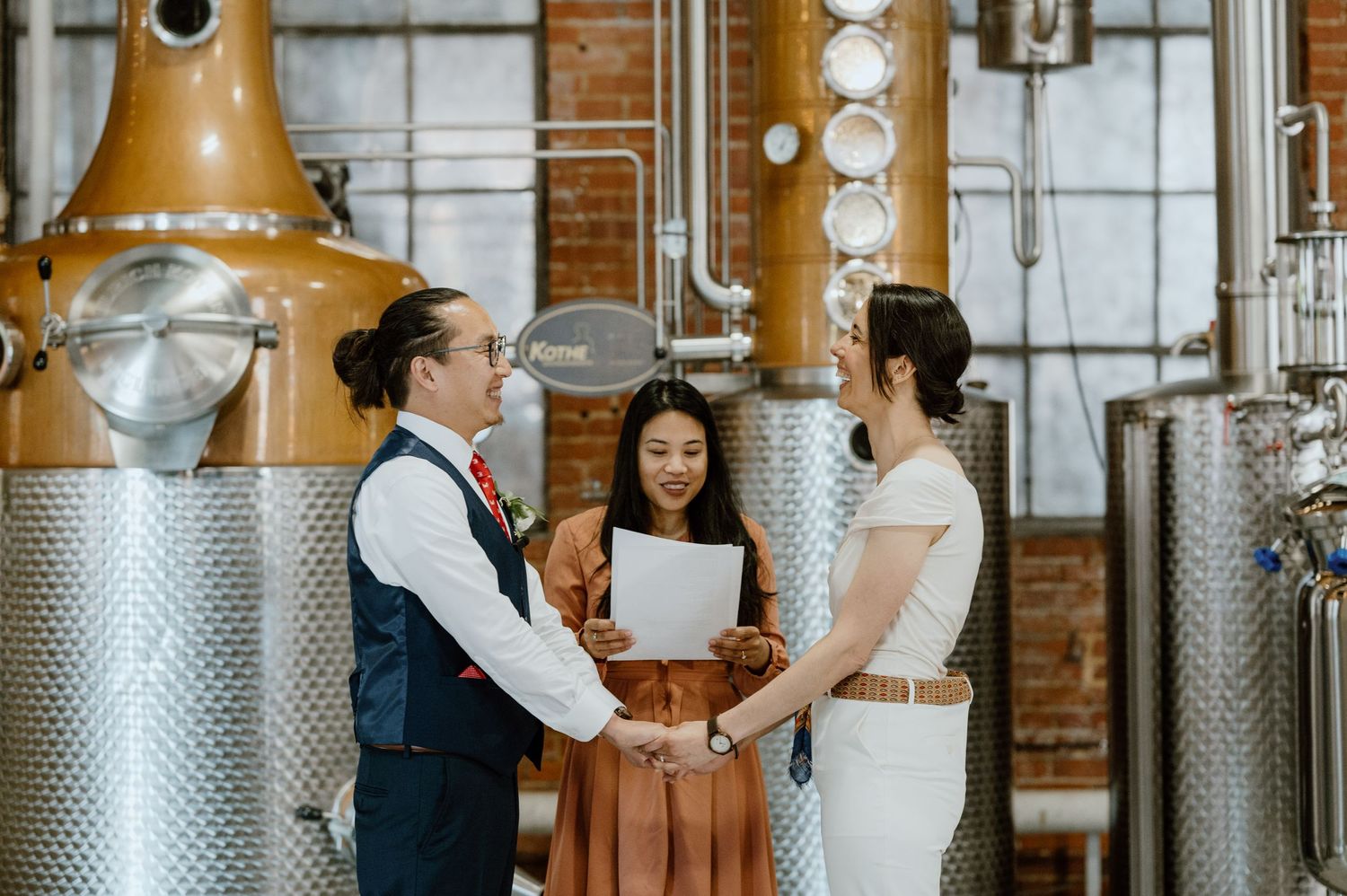 Three people participating in a ceremony in front of copper distillery stills.