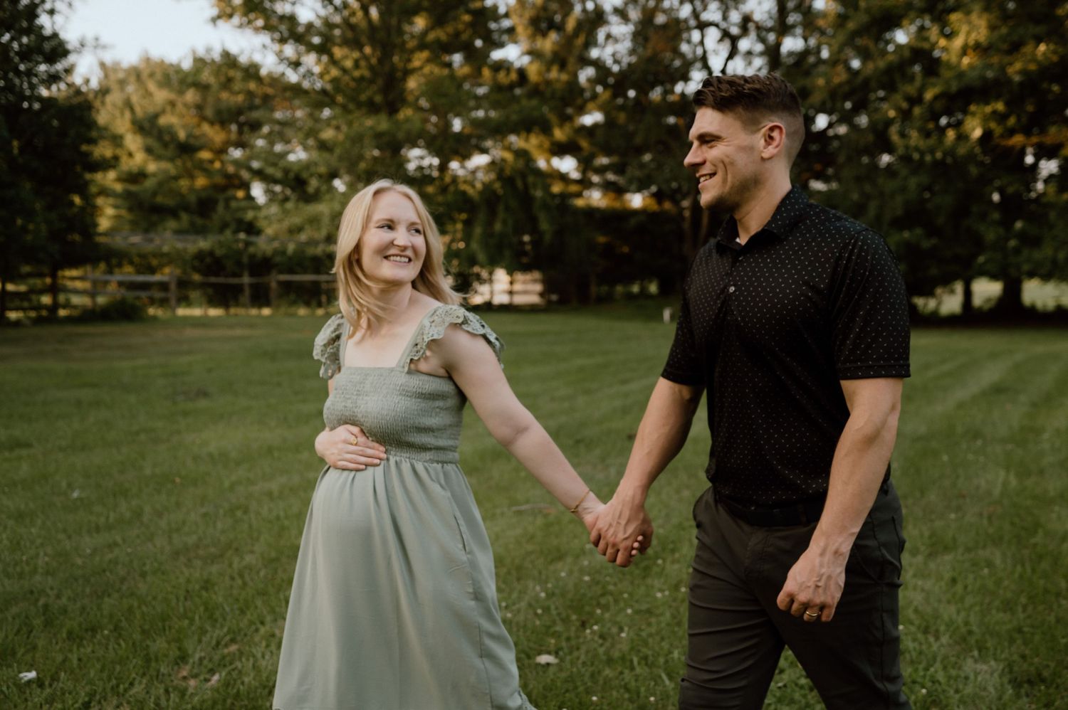 A couple holds hands while walking together in a grassy backyard garden at sunset.