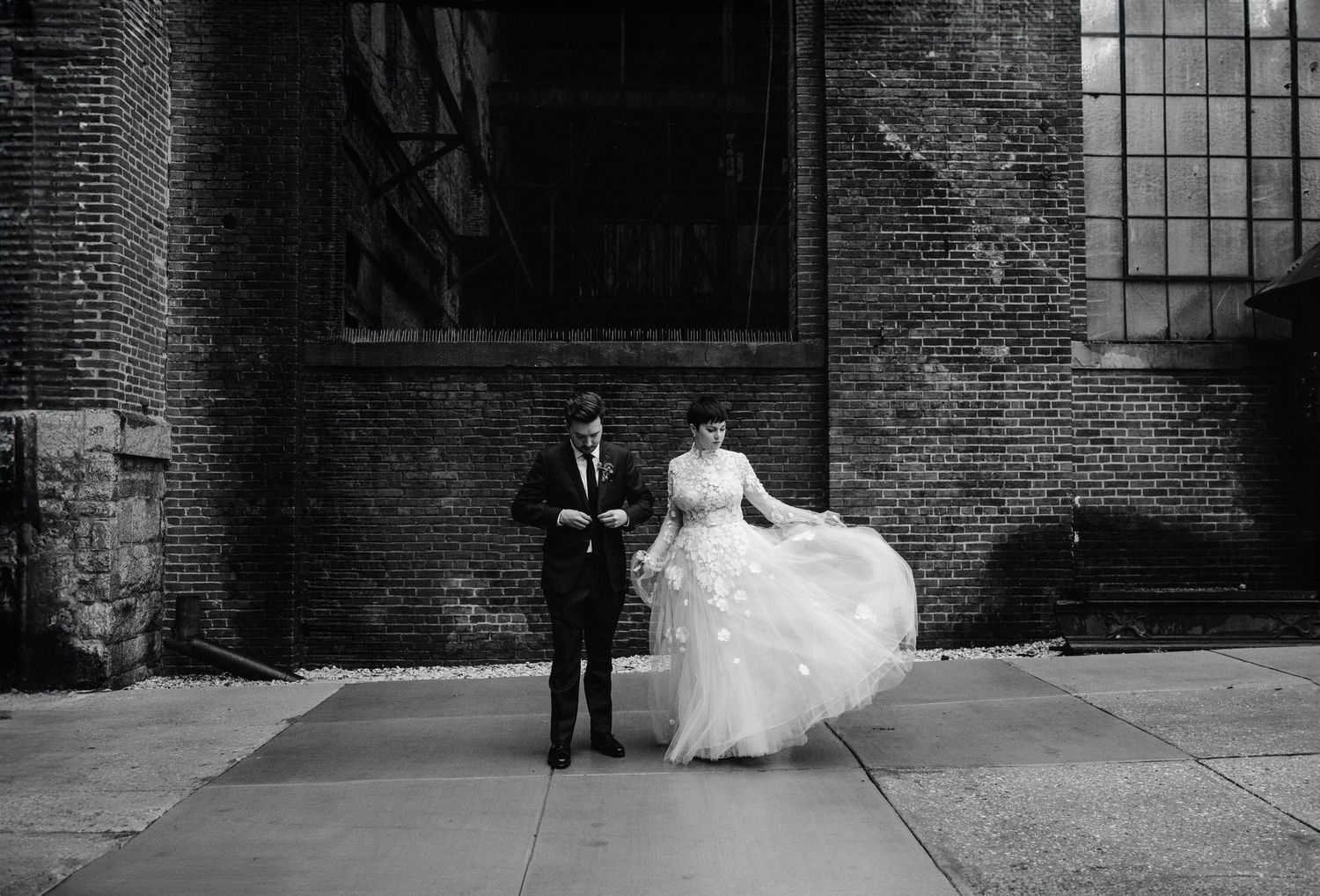 A romantic black and white wedding photo shows a fluttering dress against a historic brick building exterior.