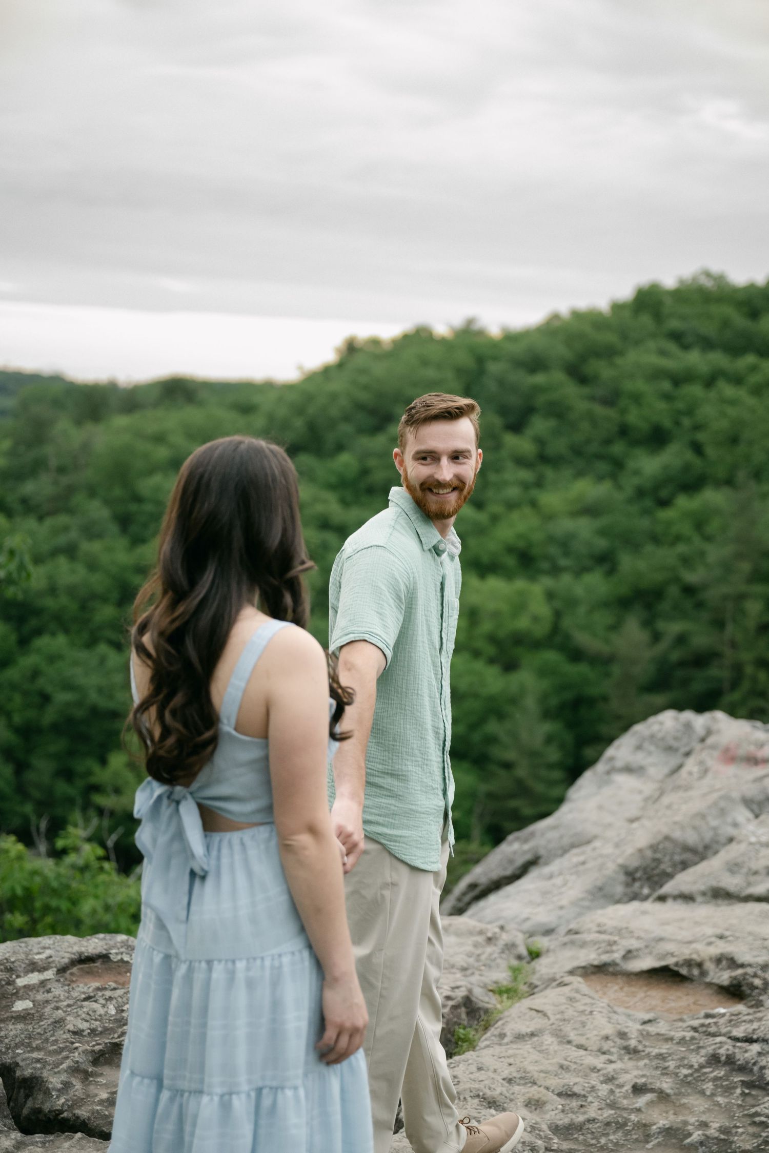 Couple sharing a playful moment on rocky overlook surrounded by green forest landscape.