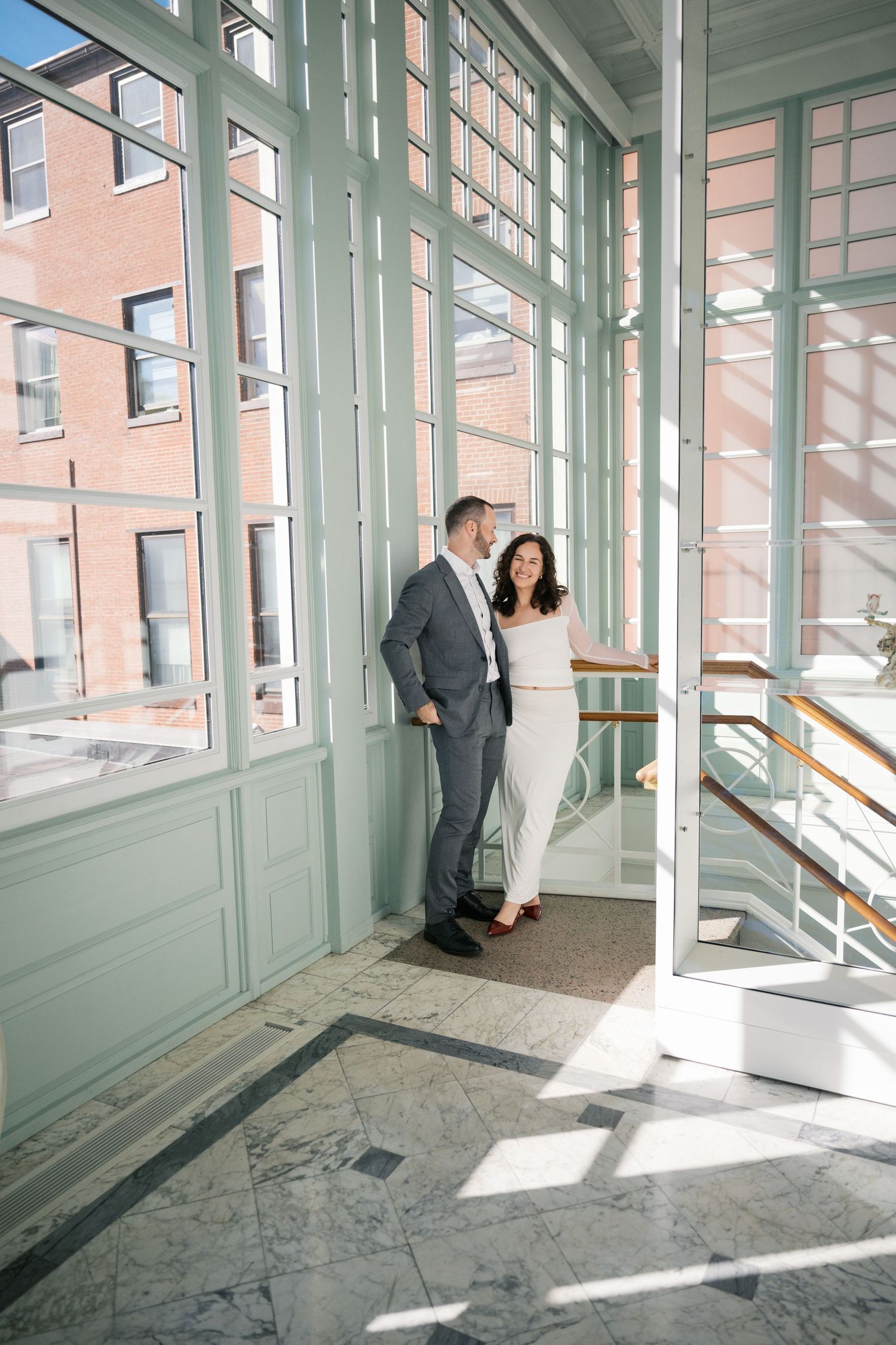 Couple in formal attire sharing a romantic moment in a sunlit glass atrium with marble floors.
