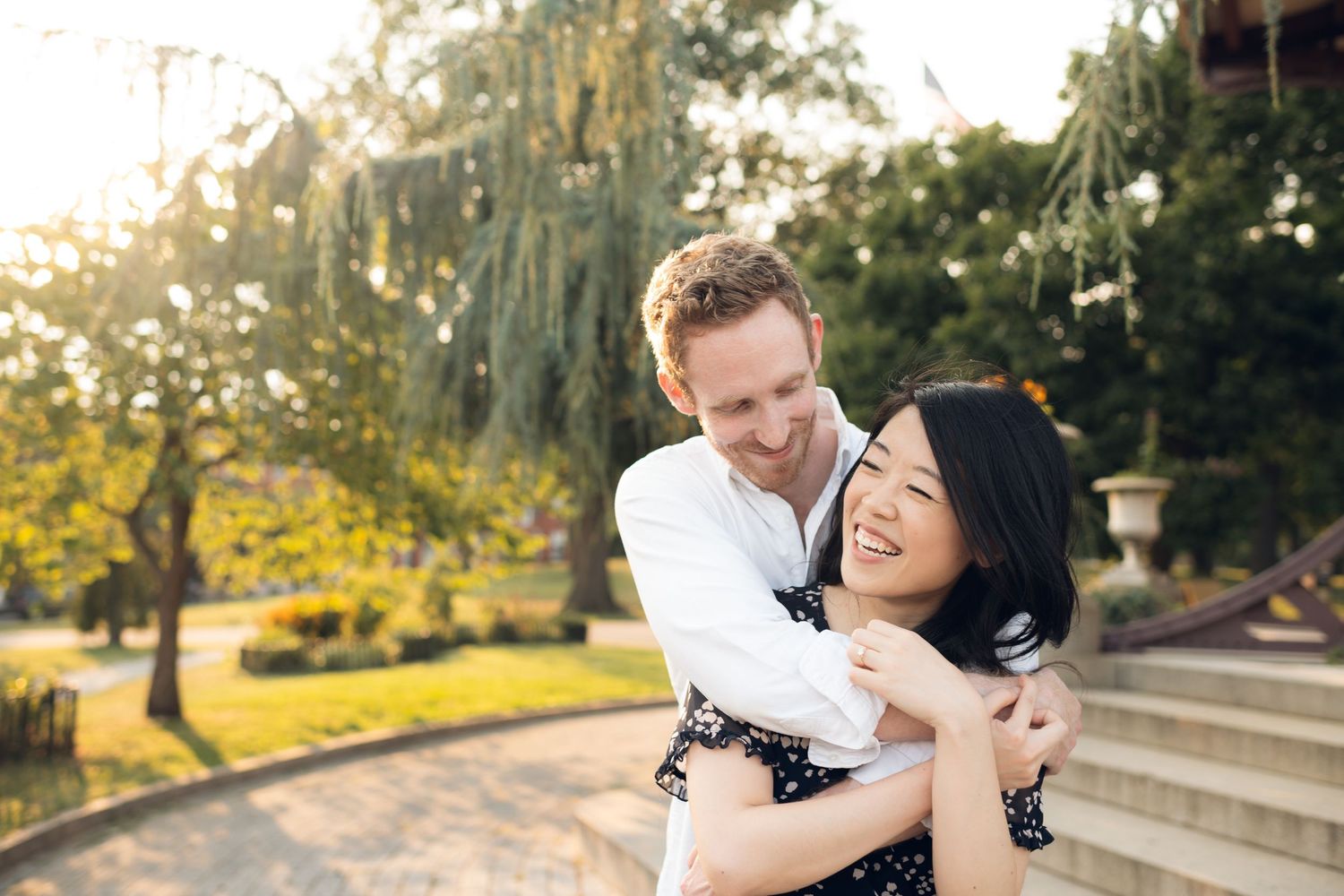 Happy interracial couple embracing outdoors in a sunlit garden, smiling joyfully during a romantic engagement photo session.