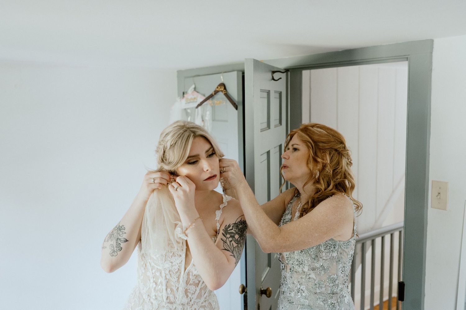 Two people in elegant dresses assist with final wedding preparations in a bright room.