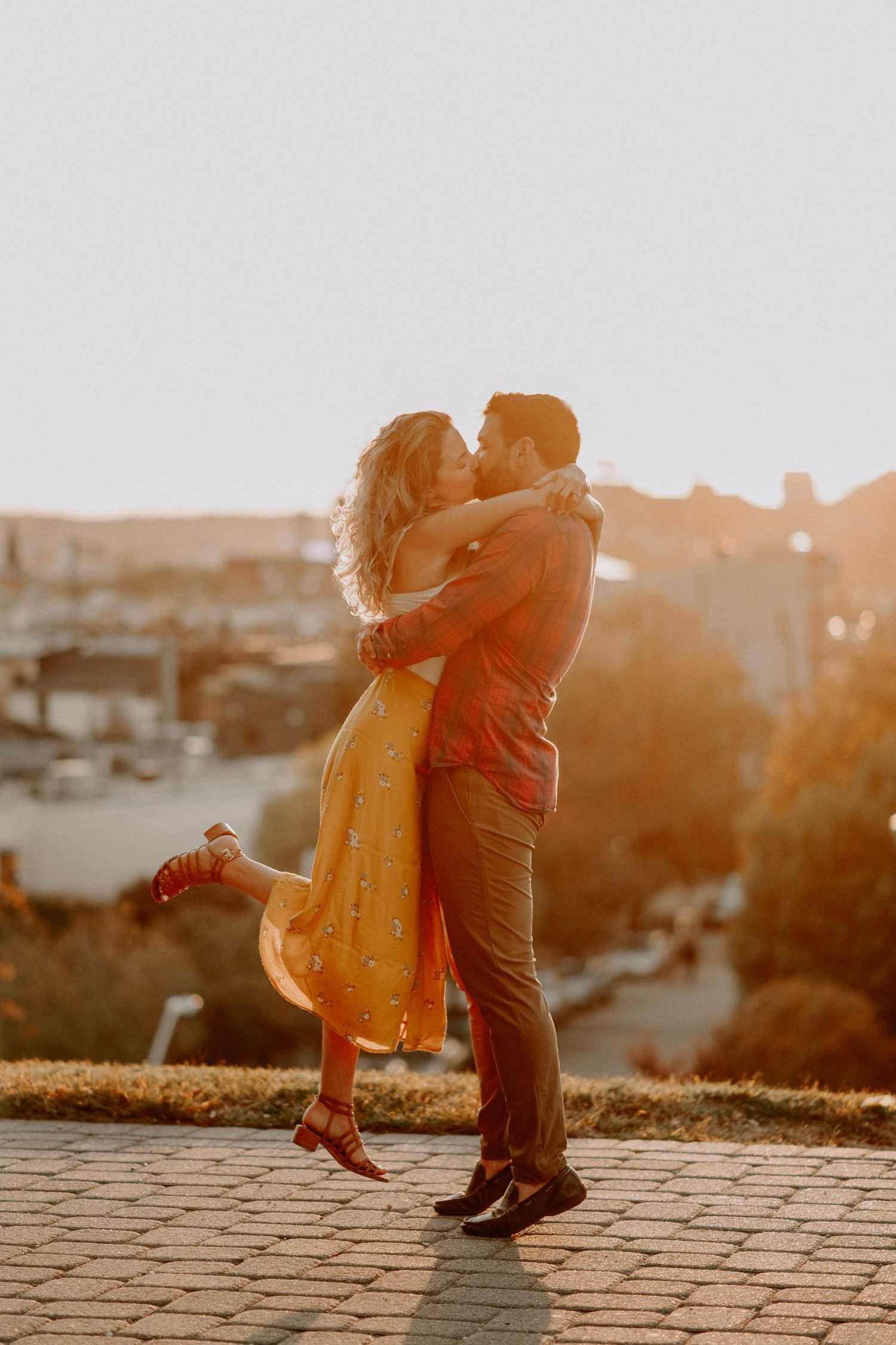 Romantic sunset embrace on cobblestone overlook with cityscape in background.