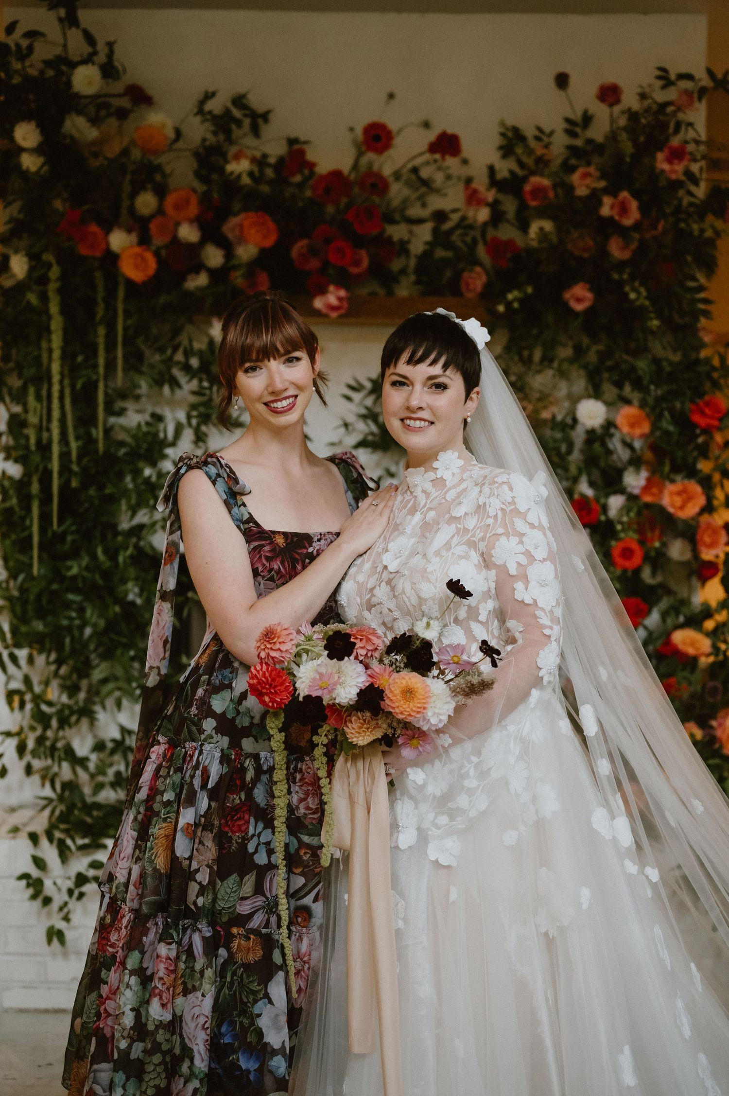 Two people pose in front of a vibrant floral arch with orange and red blooms at a wedding celebration.