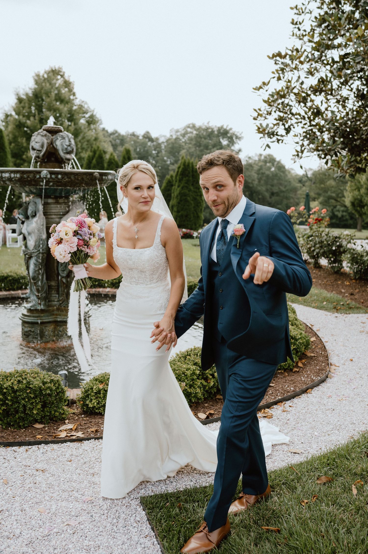 Couple celebrates on garden path near ornate fountain after wedding ceremony.