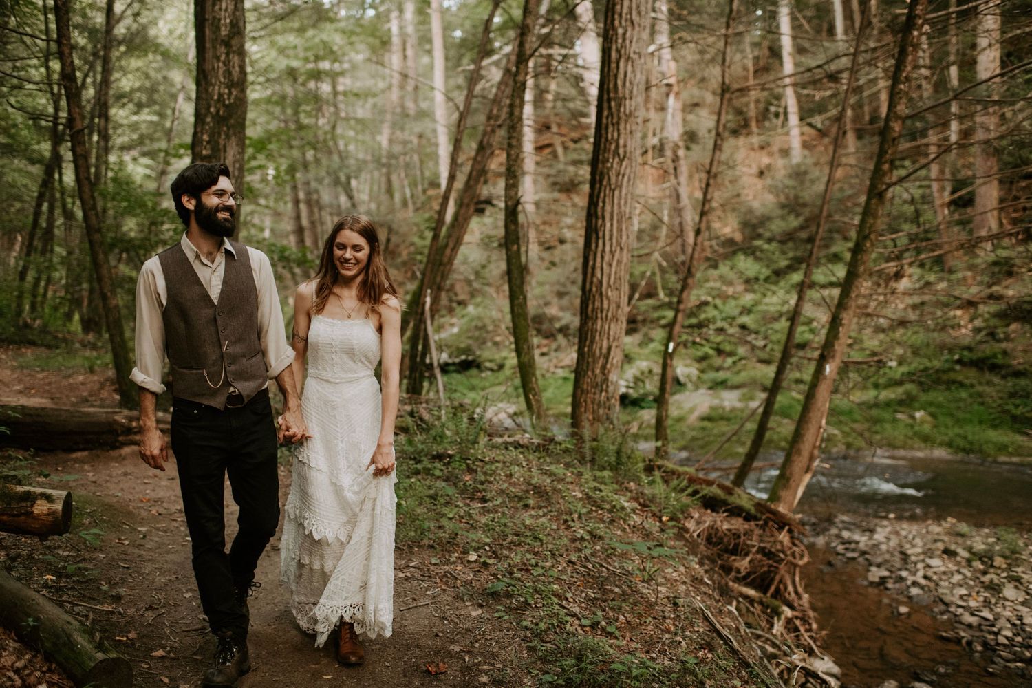 A couple walks hand in hand along a wooded trail beside a stream during their wedding day.