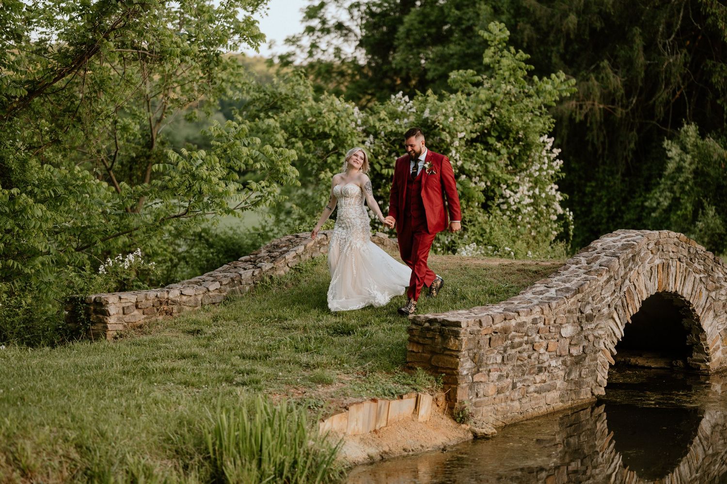 A couple in wedding attire stands on a charming stone bridge over a stream in a garden setting.