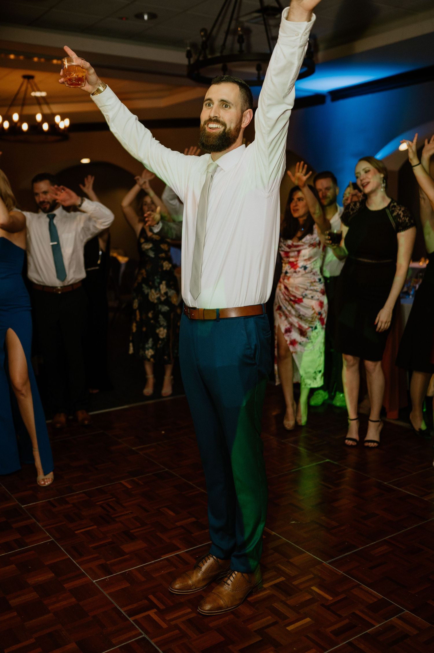 Energetic guest in white shirt and blue pants dancing enthusiastically on a wooden dance floor at a reception.