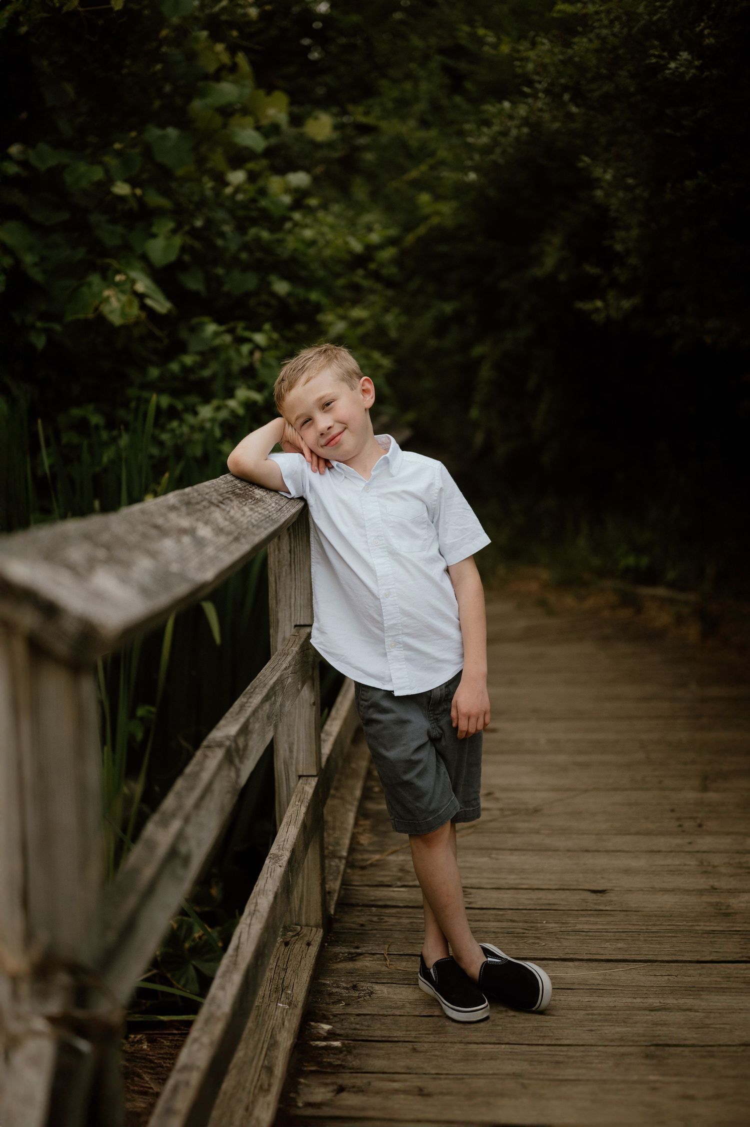 Child in white shirt leans against wooden railing of nature boardwalk surrounded by greenery.