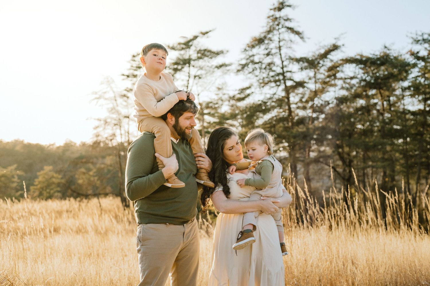 Family stands together in golden field at sunset with trees in background.