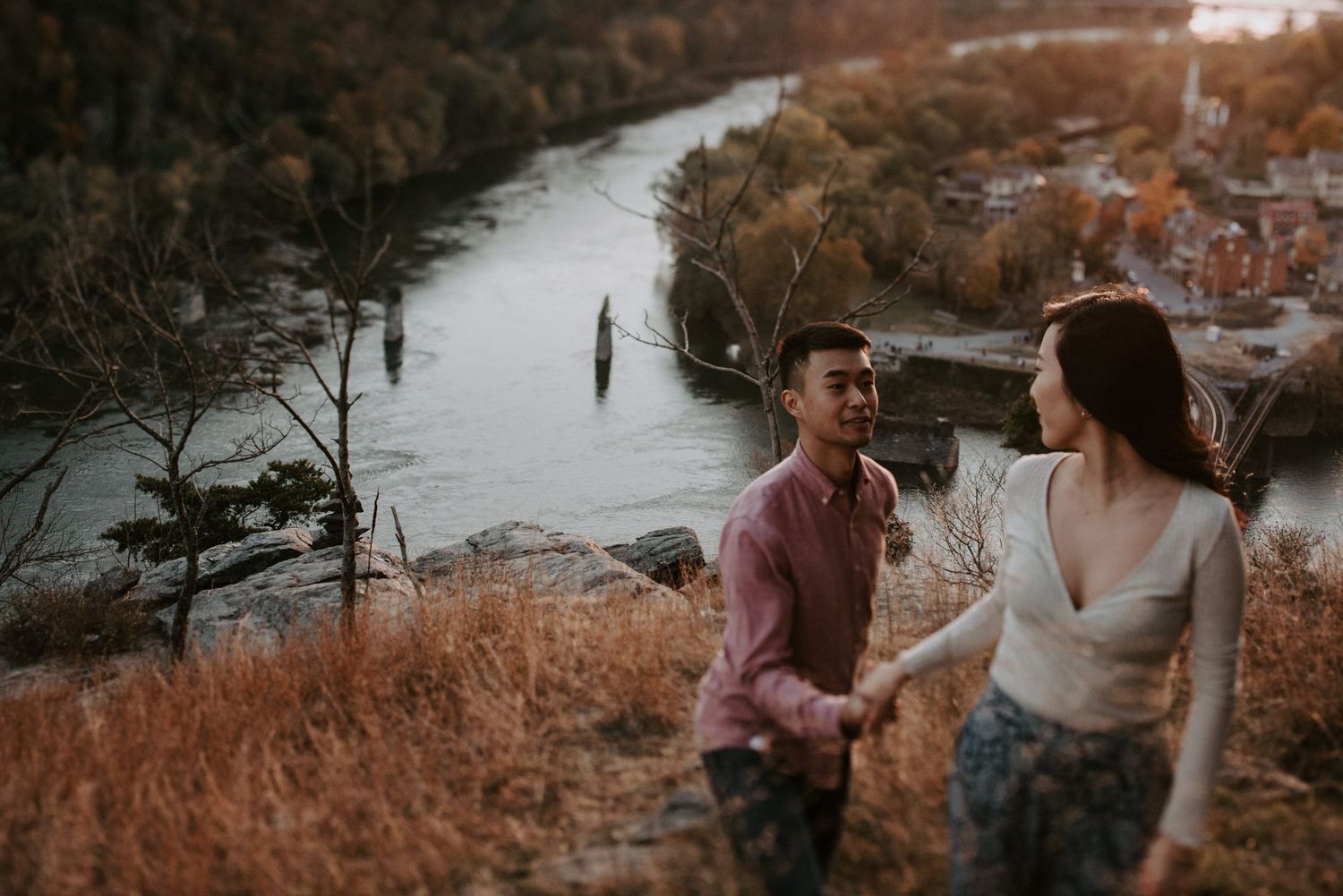 Couple sharing romantic moment on cliff edge with river valley vista behind them.
