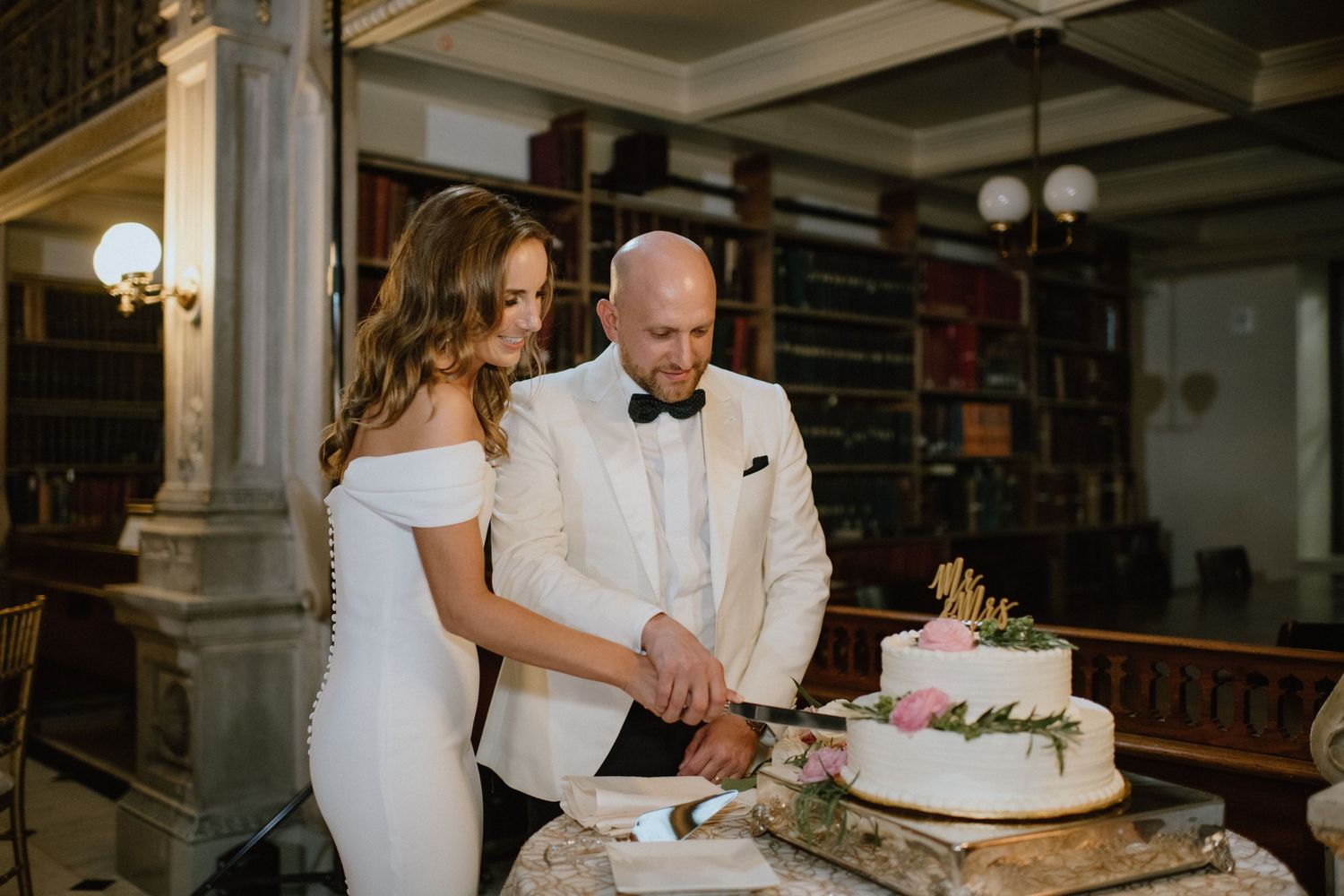 Couple cuts their wedding cake together in elegant library setting with vintage bookshelves.