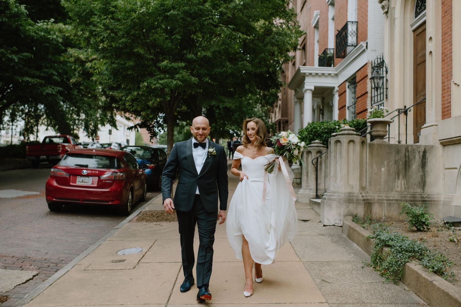 Wedding couple walking together on a city sidewalk past parked cars and historic buildings.