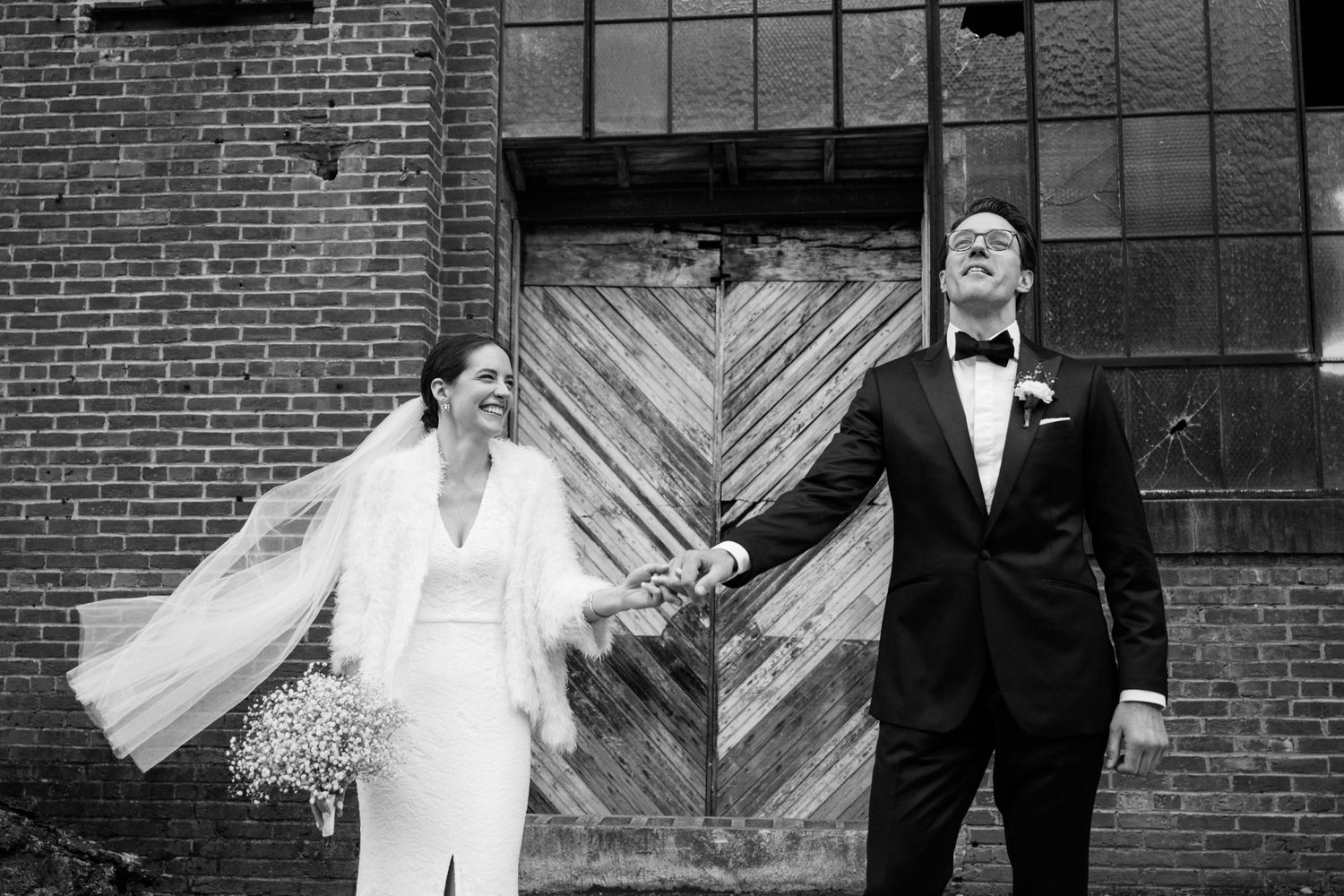 Artistic black and white wedding photo against industrial brick building with wooden door.