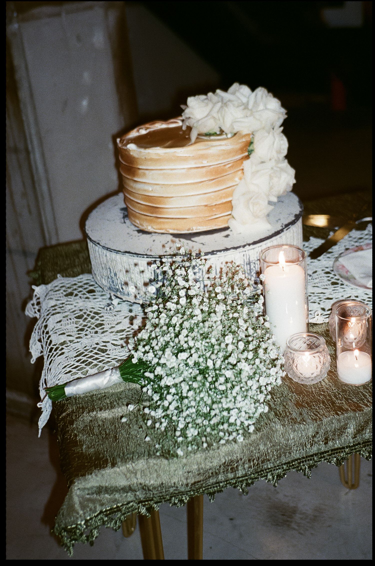 Two-tier rustic wedding cake with white flowers and baby's breath on a velvet-covered table with glowing candles.