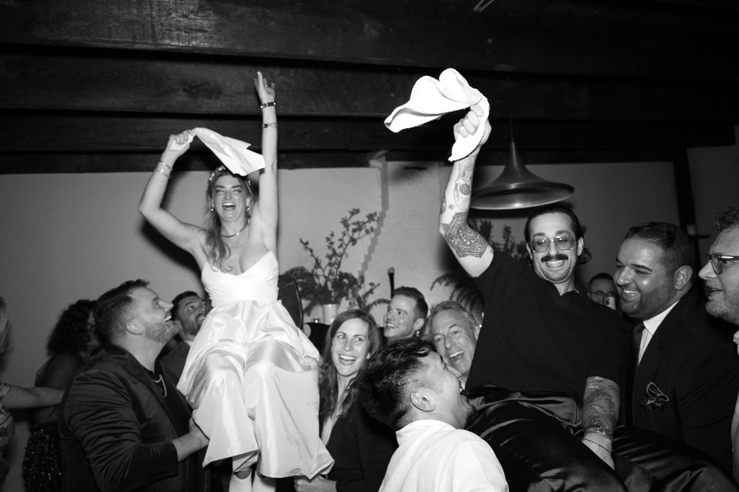 Celebration at an indoor wedding reception shows people dancing and waving white napkins in a festive black and white photo.