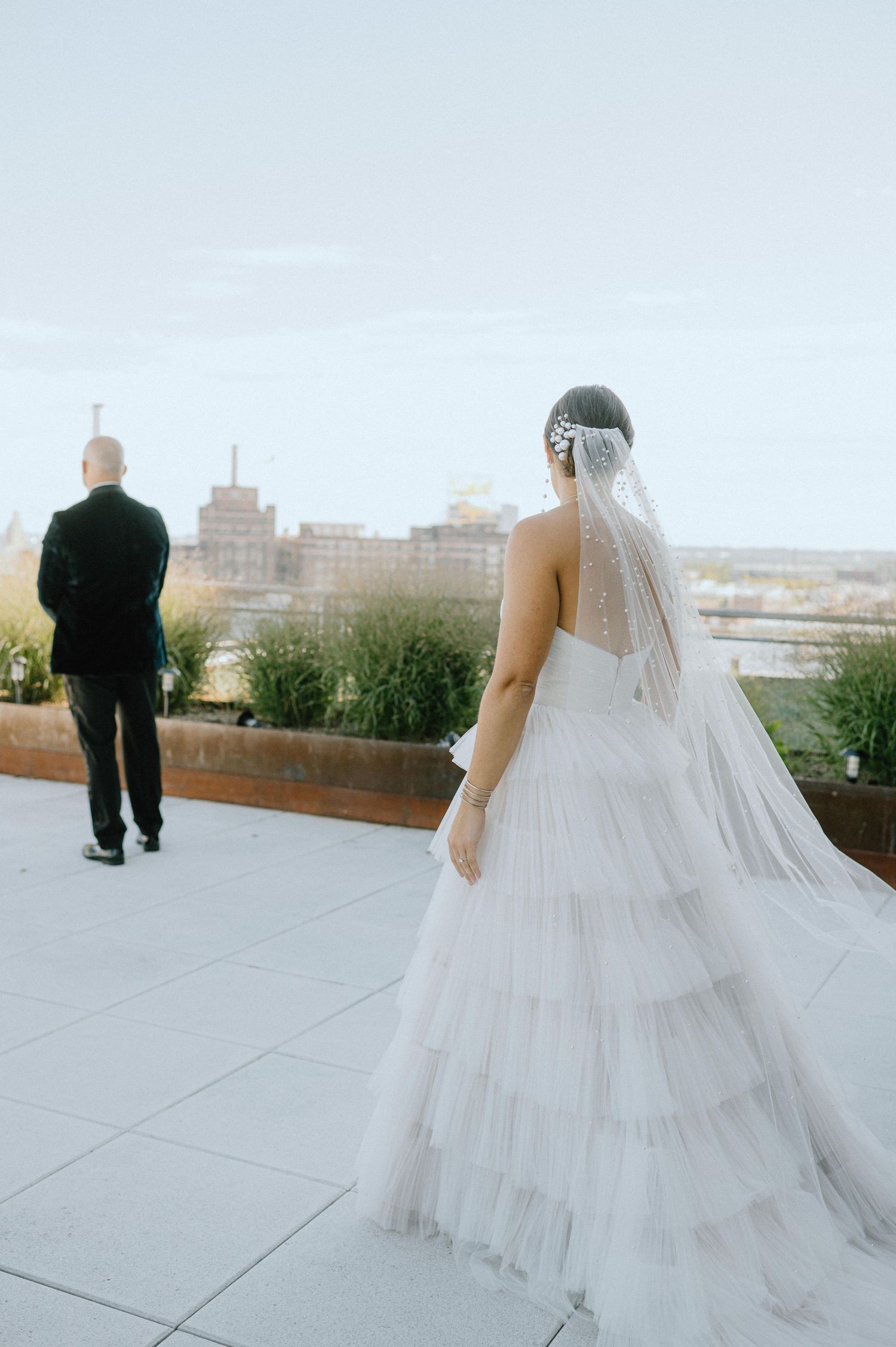 A bride in a layered tulle gown and veil stands on a rooftop terrace overlooking a city.