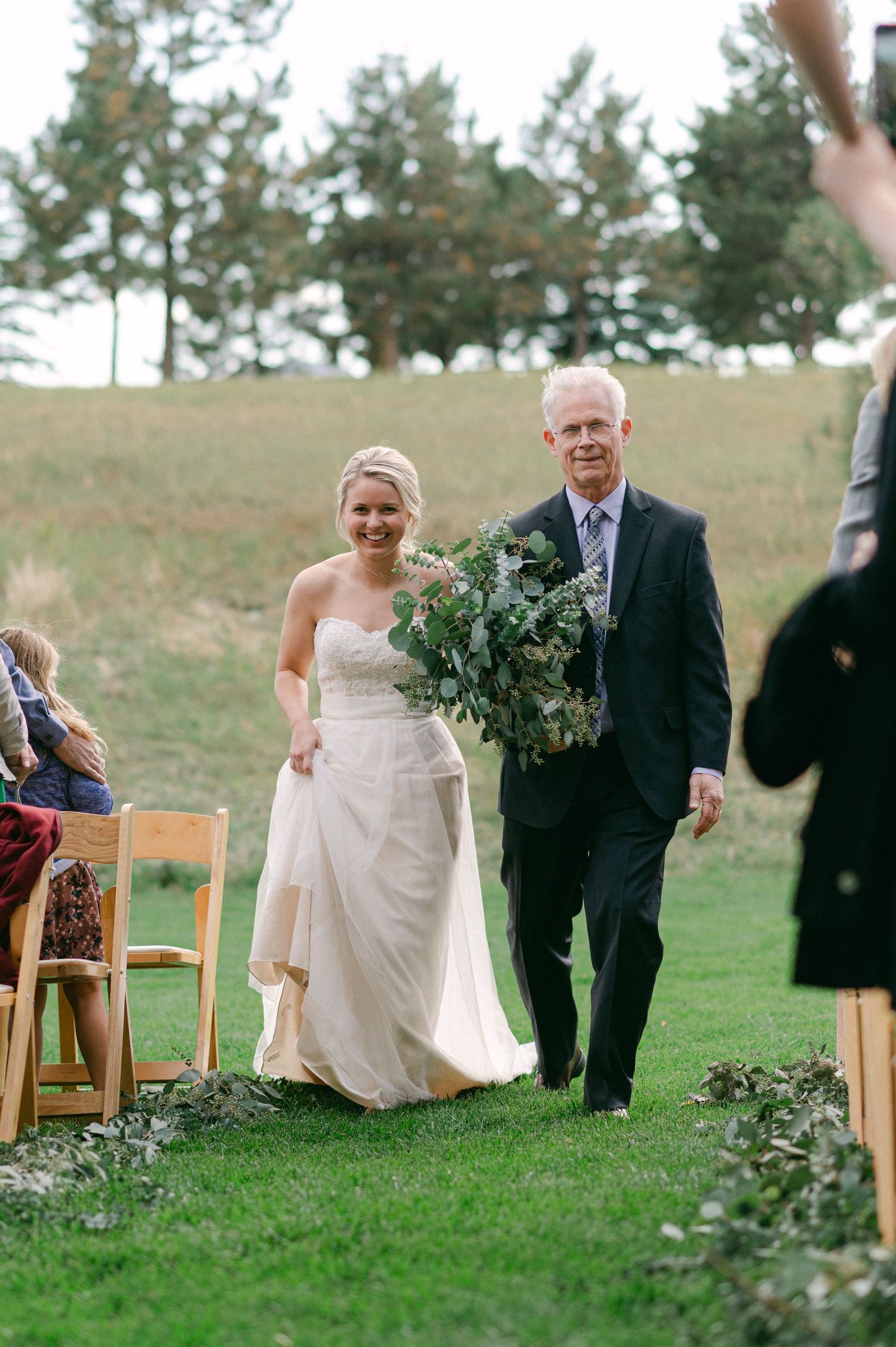 Wedding processional on a grassy hillside with wooden chairs lining the aisle.