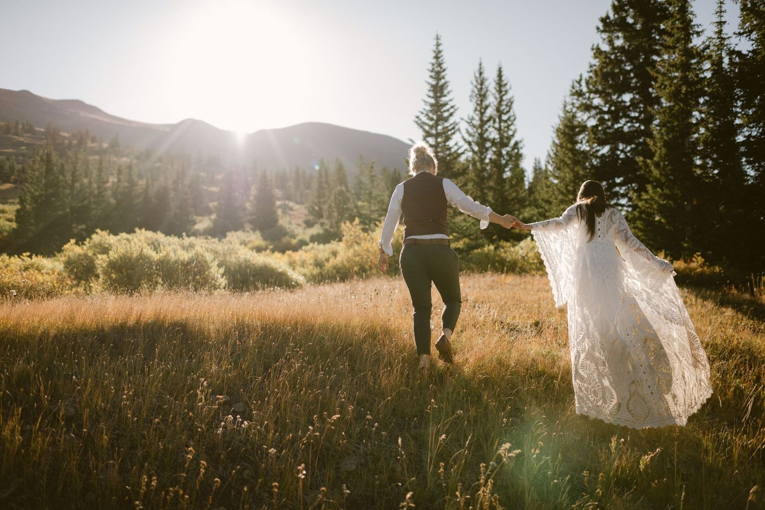 Two people walking through tall grass with mountains and pine trees in the background.