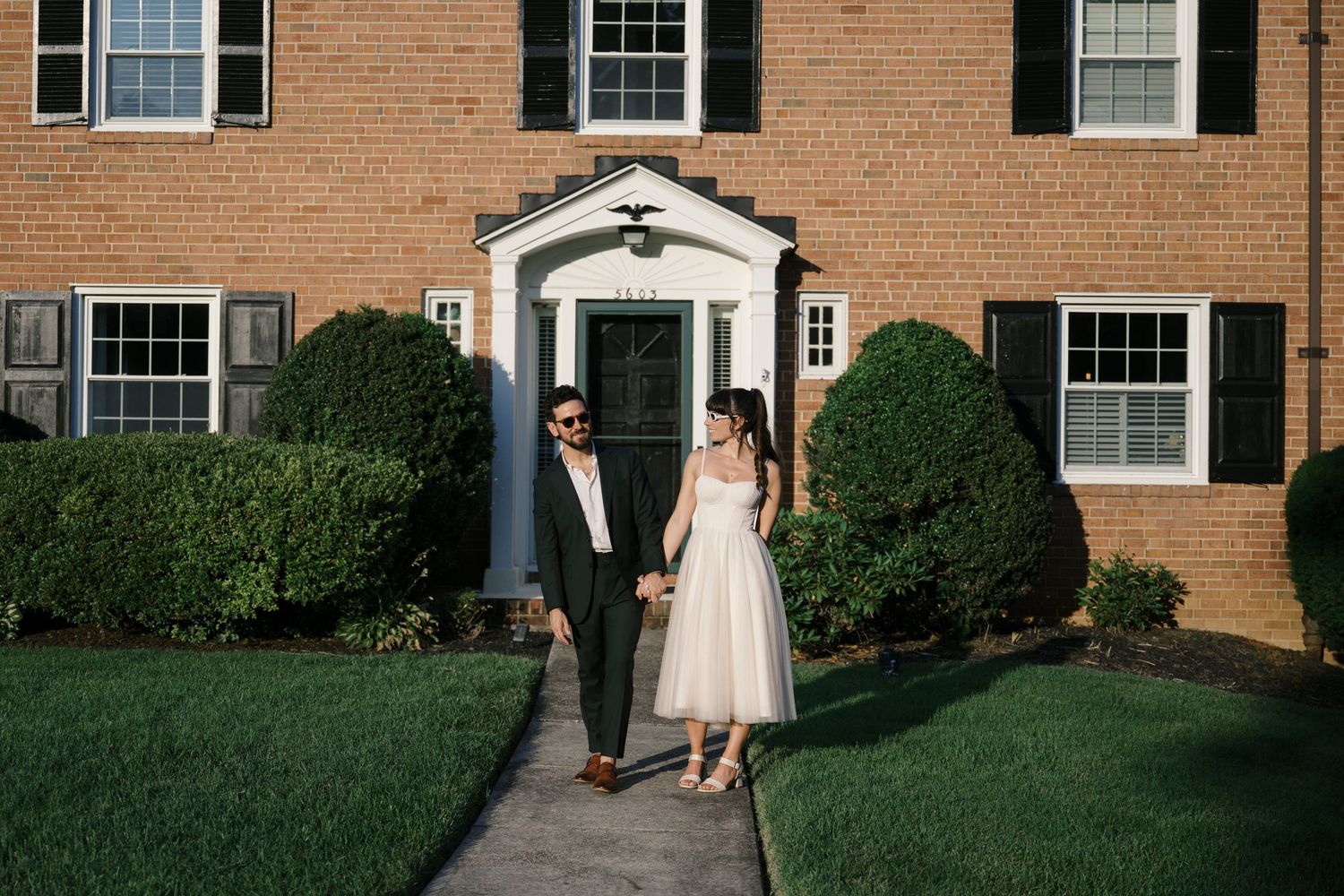 A couple walks along a brick residential building with white trim windows on a sunny day.