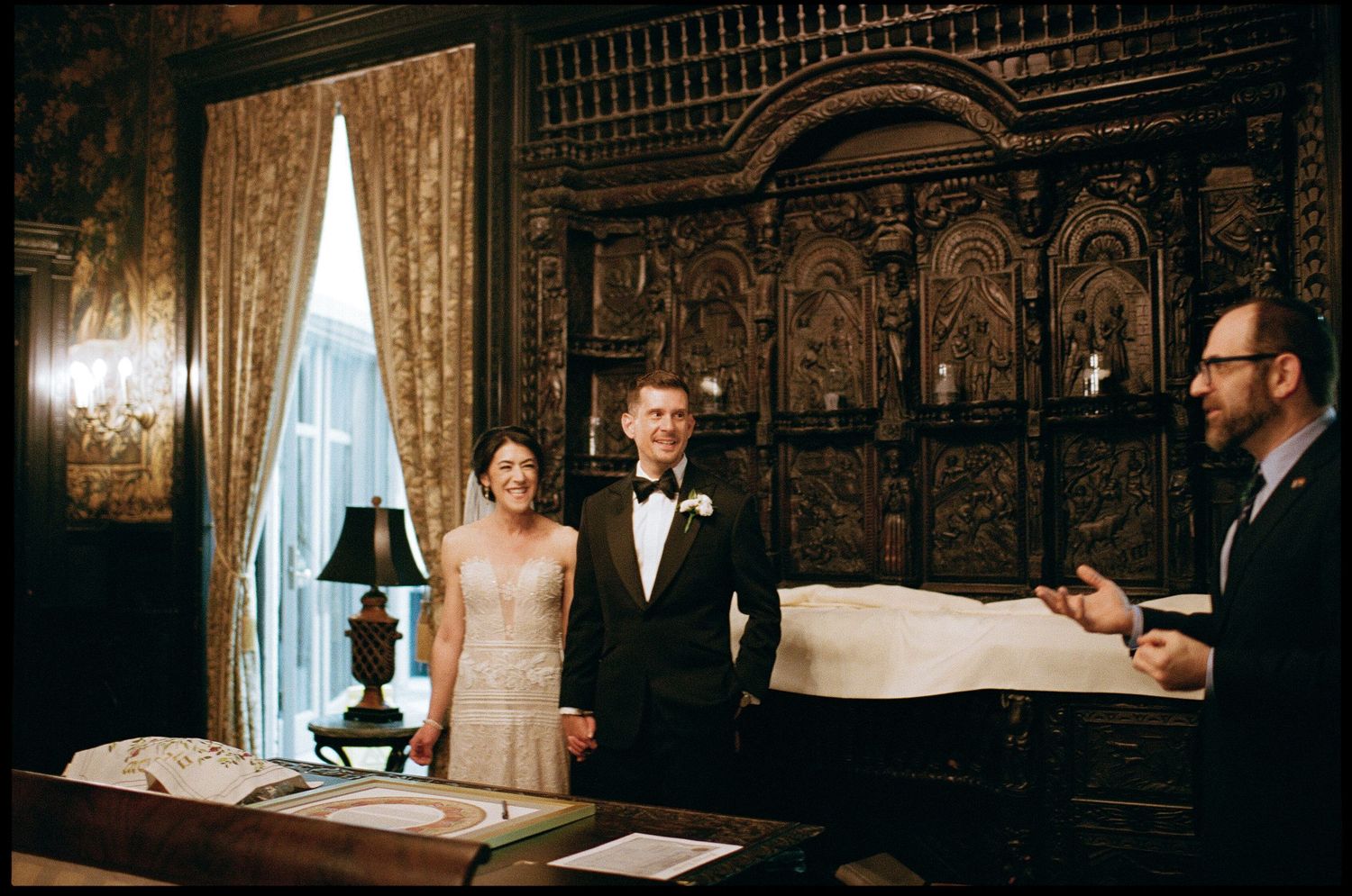 A couple stands in an ornate antique room with carved wood paneling and a large window while speaking with another person.