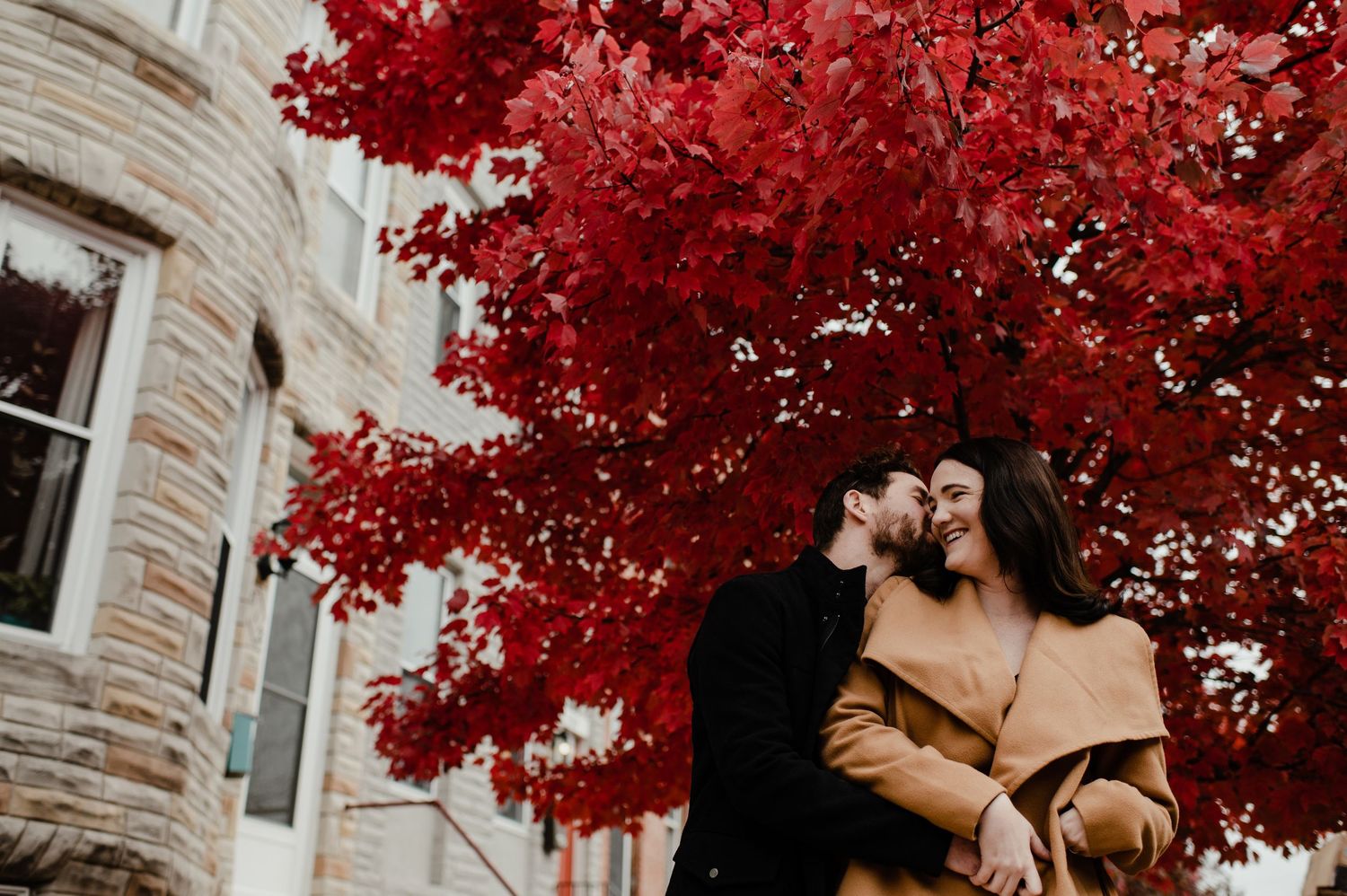 Couple embracing and smiling under a vibrant red autumn tree beside a stone building.