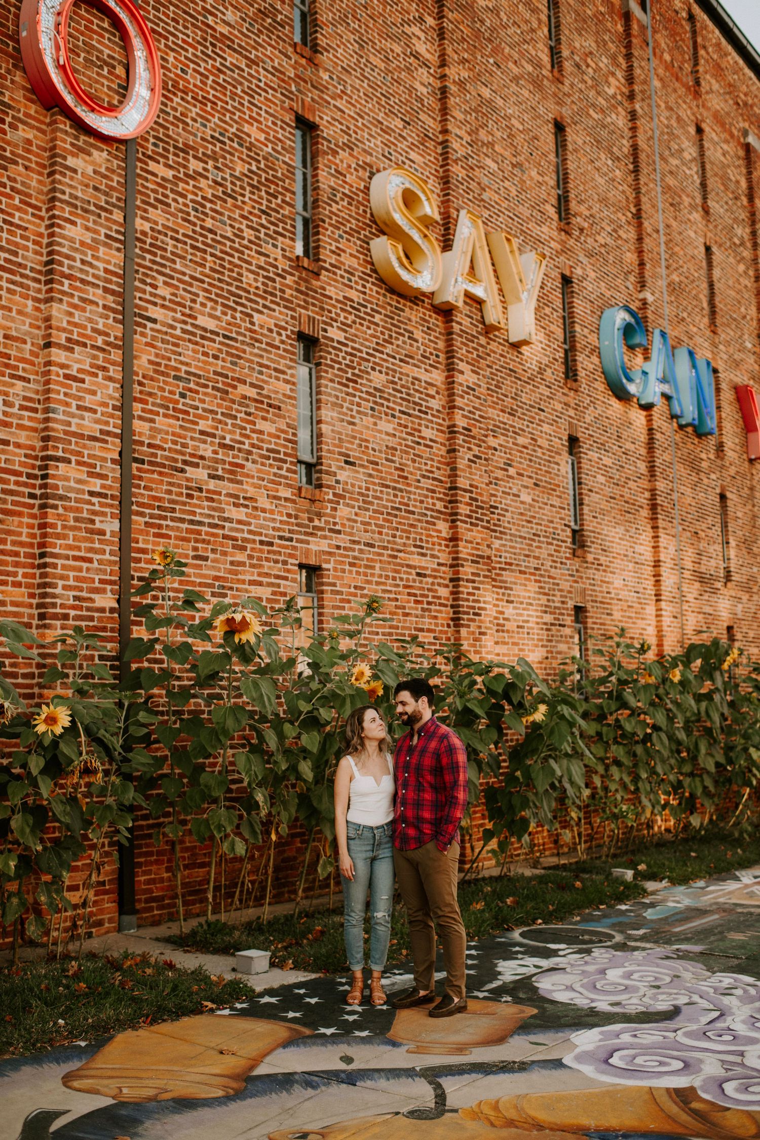 Couple stands before a brick wall with SAY CAN signage surrounded by sunflowers and street art in an urban setting.