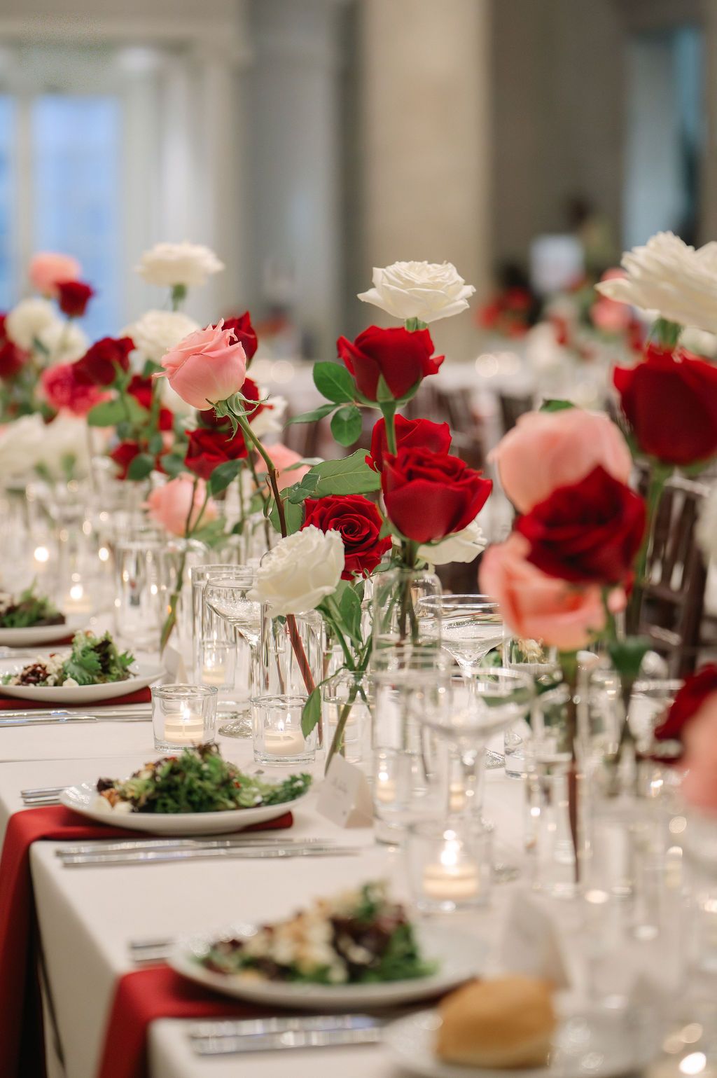Elegant wedding reception table decorated with red and pink roses in glass vases alongside white place settings and glowing candles.