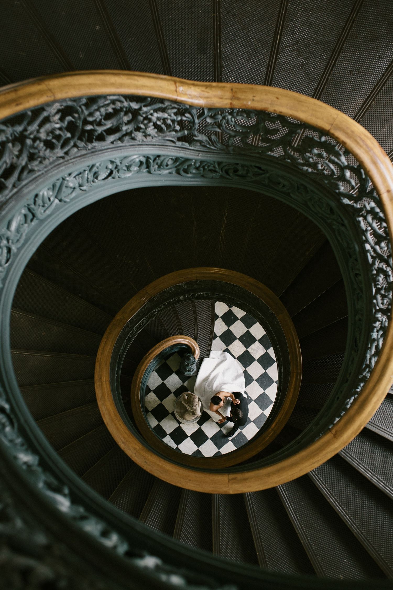 Spiral staircase with ornate railing viewed from above showing checkered floor pattern below.