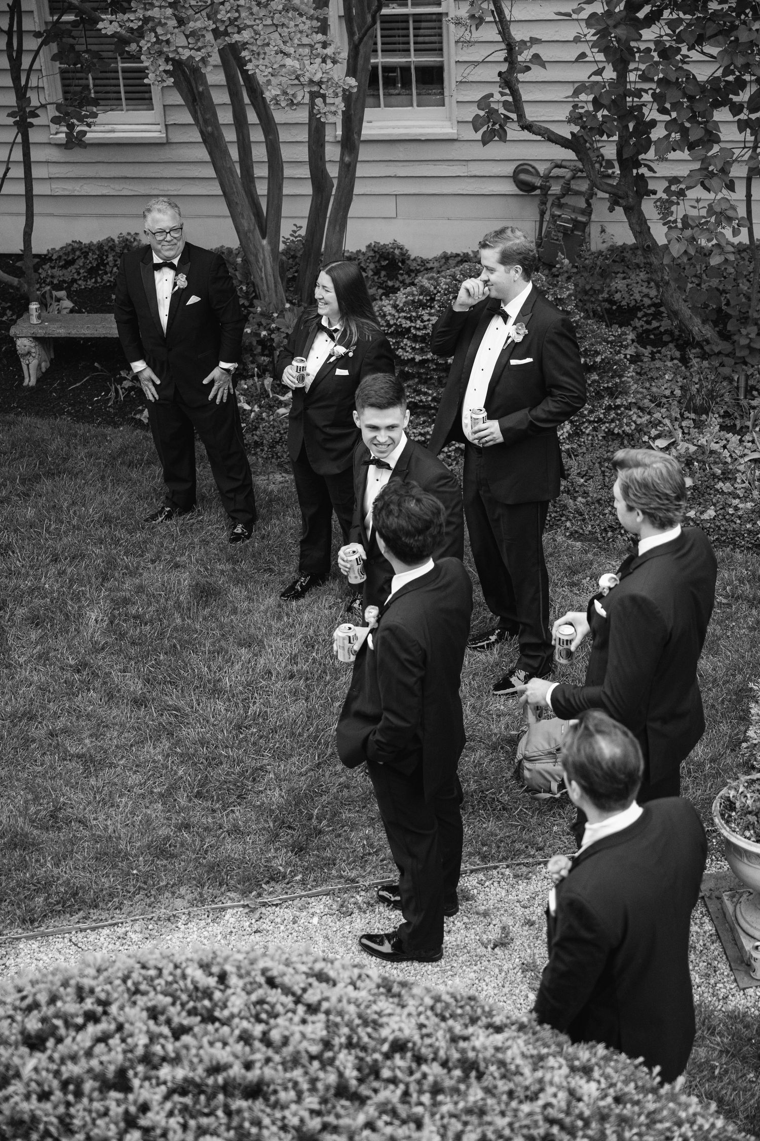 Black and white photo of groomsmen in tuxedos socializing outdoors at a wedding reception.