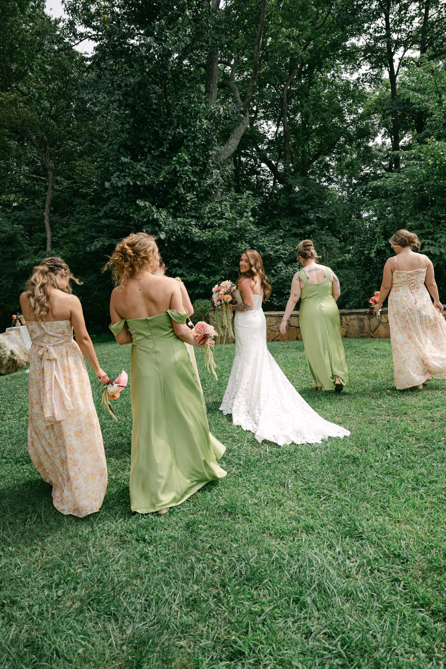 Bride in white gown walks with bridesmaids in green and floral dresses across lush garden lawn.
