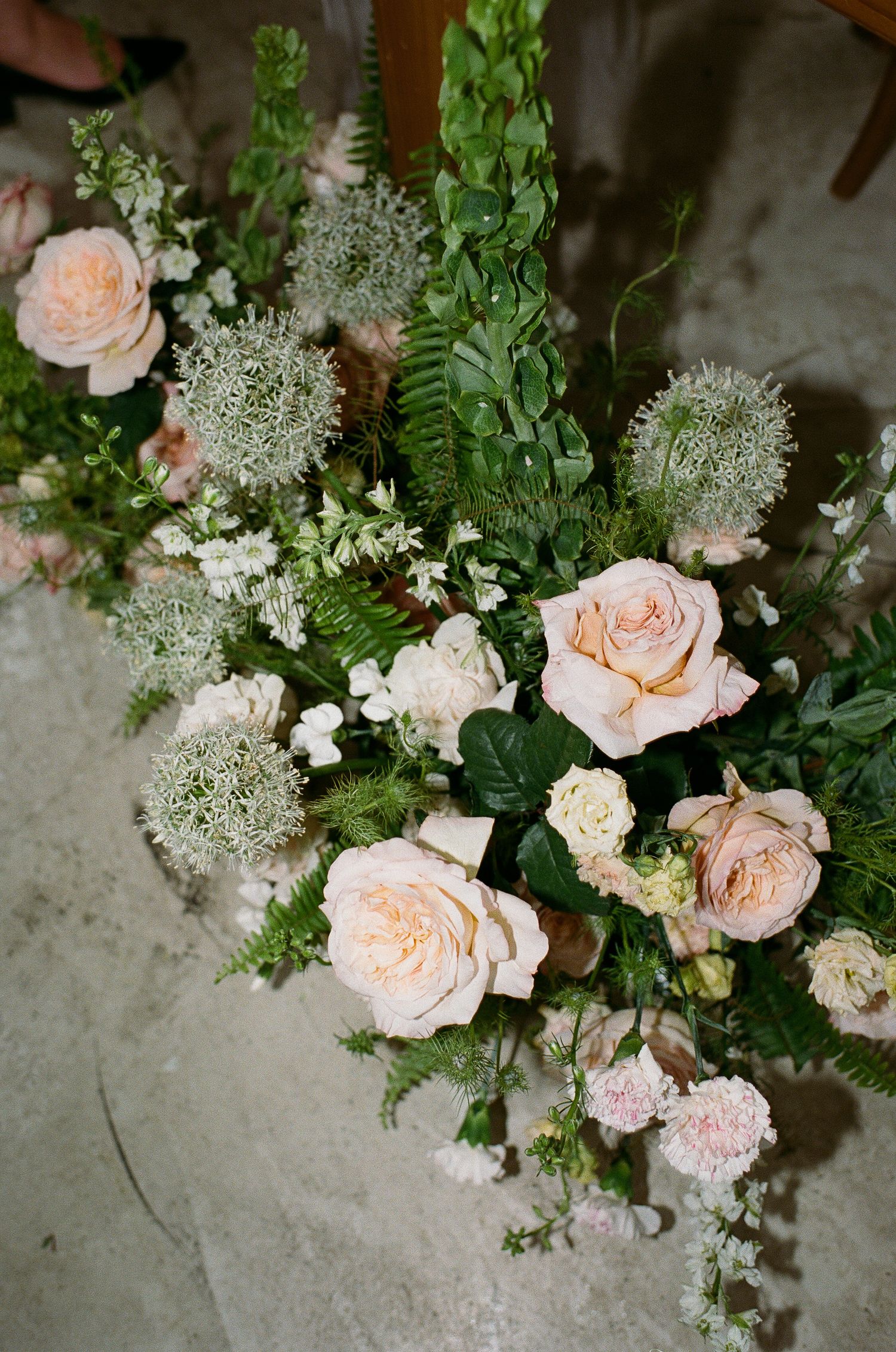 Elegant floral arrangement with blush roses, ferns, and white blooms on a stone surface for a wedding.