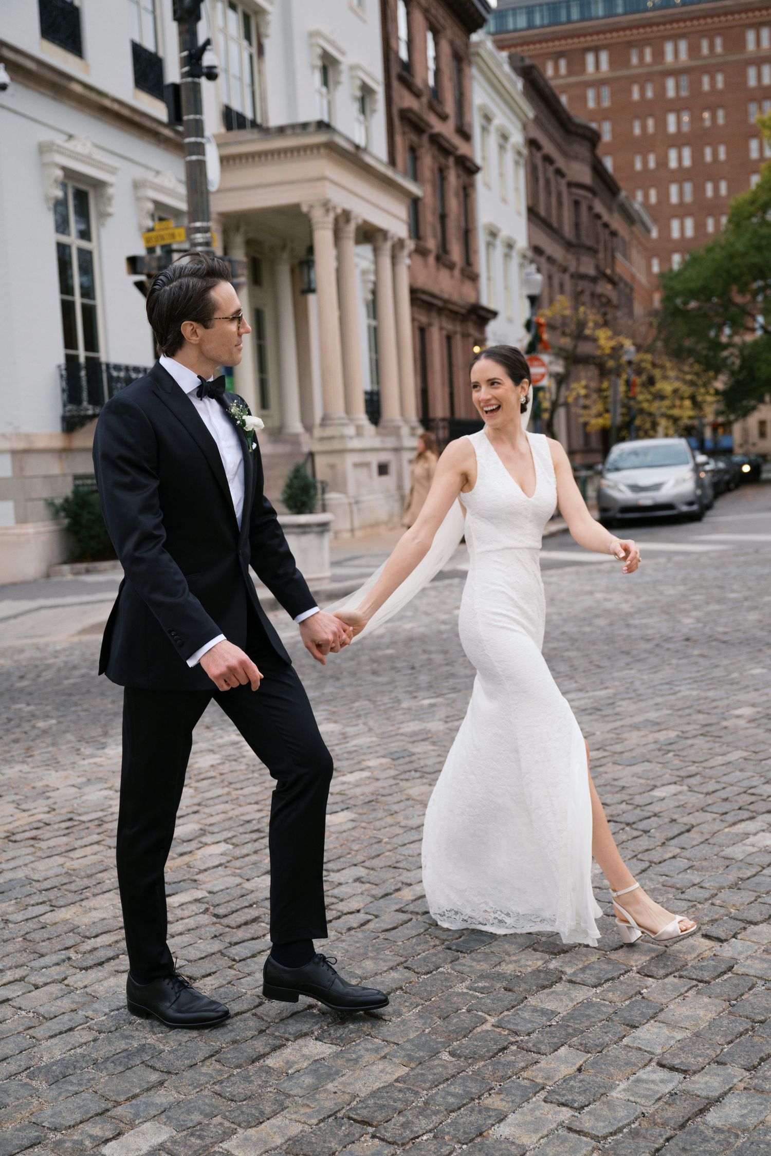 Elegant bride and groom hold hands while walking on cobblestone street in urban setting.