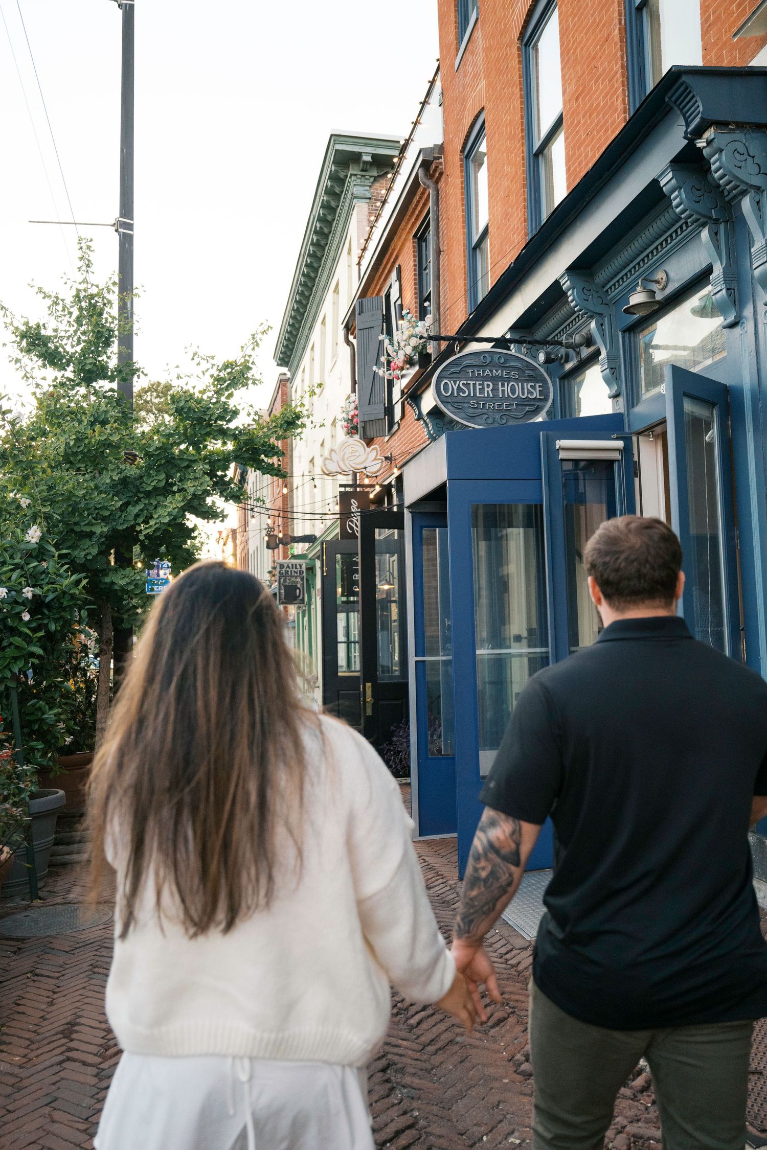 A couple walks down a historic brick-paved street past storefronts and shops with decorative architecture.