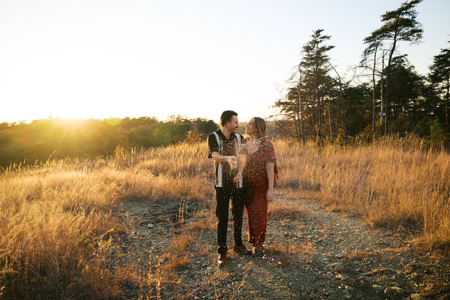 A couple shares an intimate moment on a dirt path surrounded by golden grass and evergreen trees at sunset.