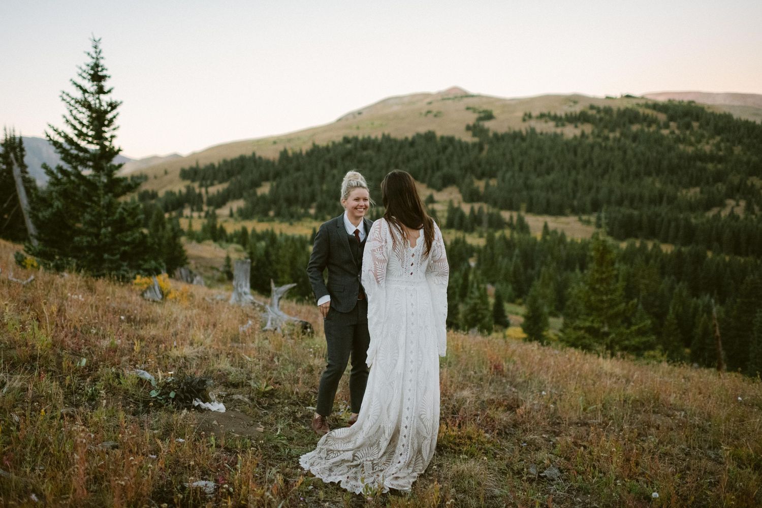 Couple stands on mountainside meadow with forest and peaks in background during golden hour.