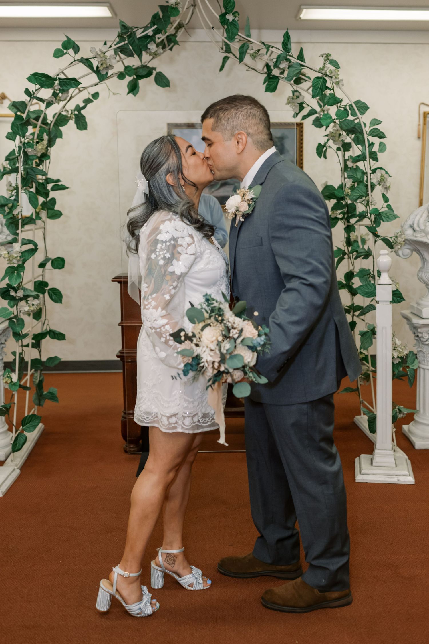 A romantic wedding moment under a green ivy arch with a bride in a short lace dress and groom in gray suit sharing a kiss.