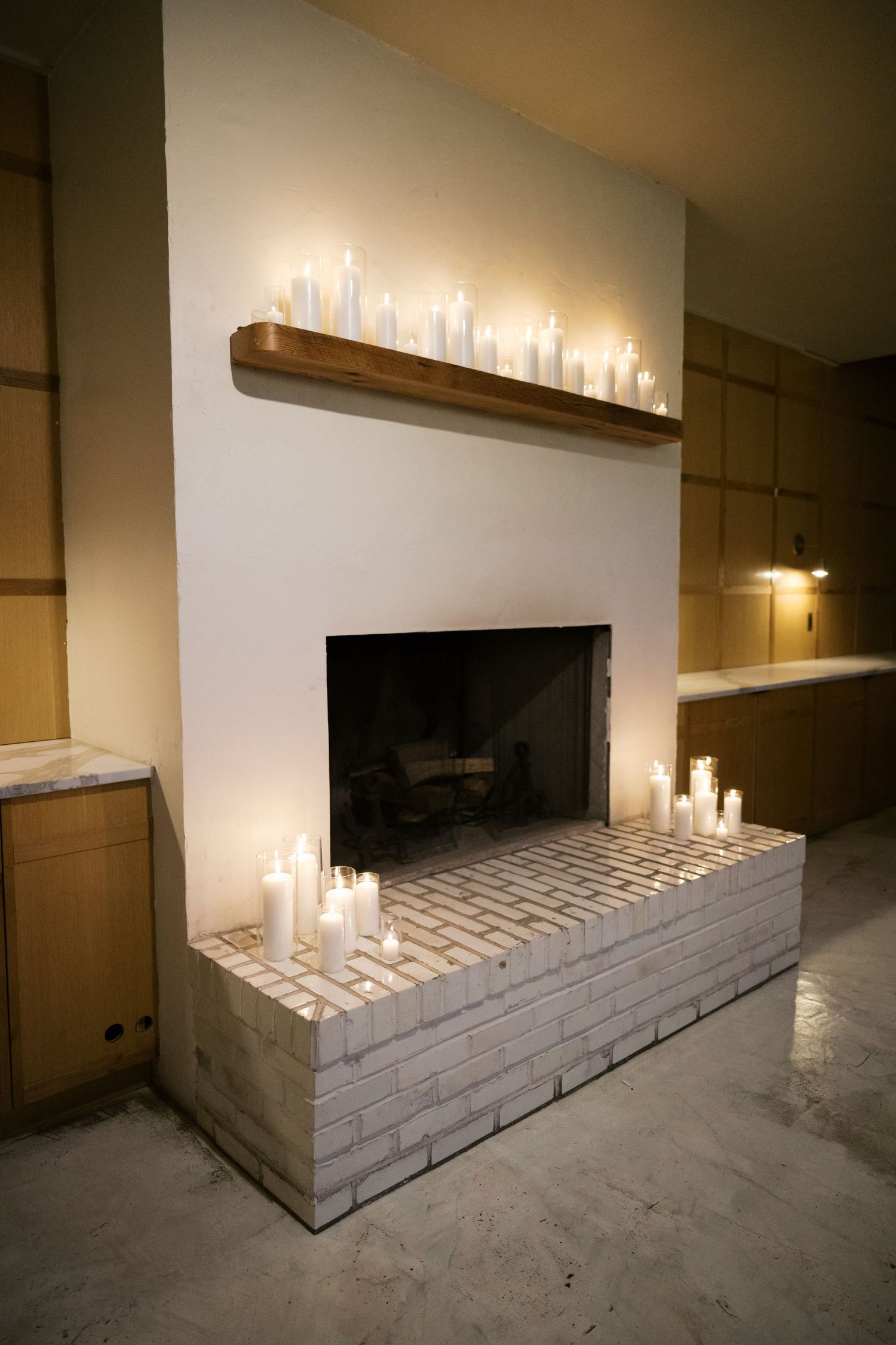 Modern fireplace decorated with numerous white pillar candles on a brick hearth and floating wooden shelf.