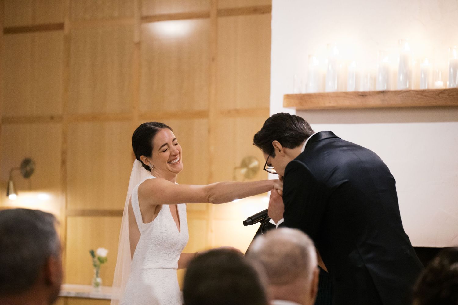 Two figures in wedding attire share a tender laugh together at a ceremony in an indoor venue.