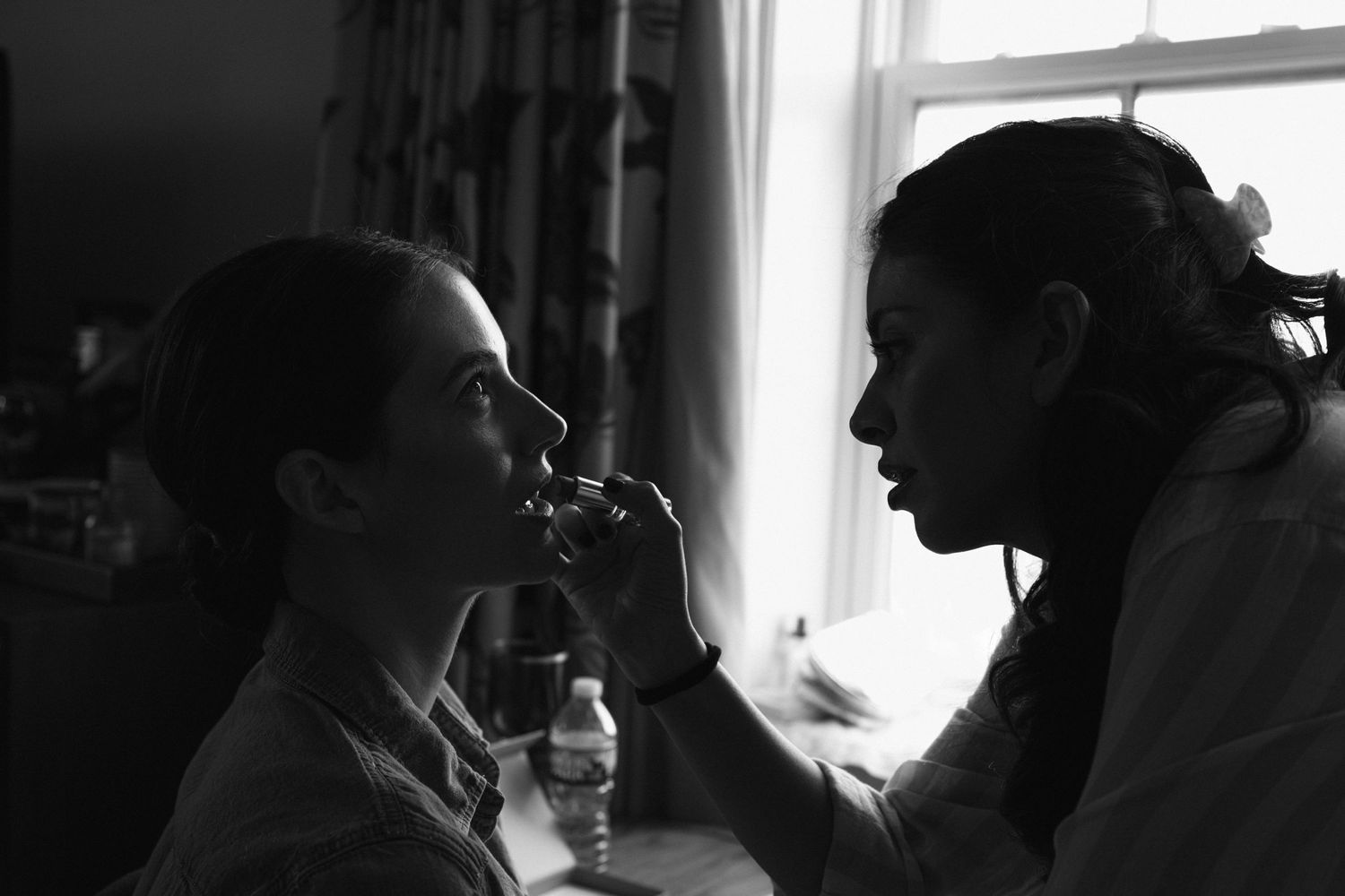 Black and white artistic portrait of a makeup artist applying lipstick to a model in natural window light.