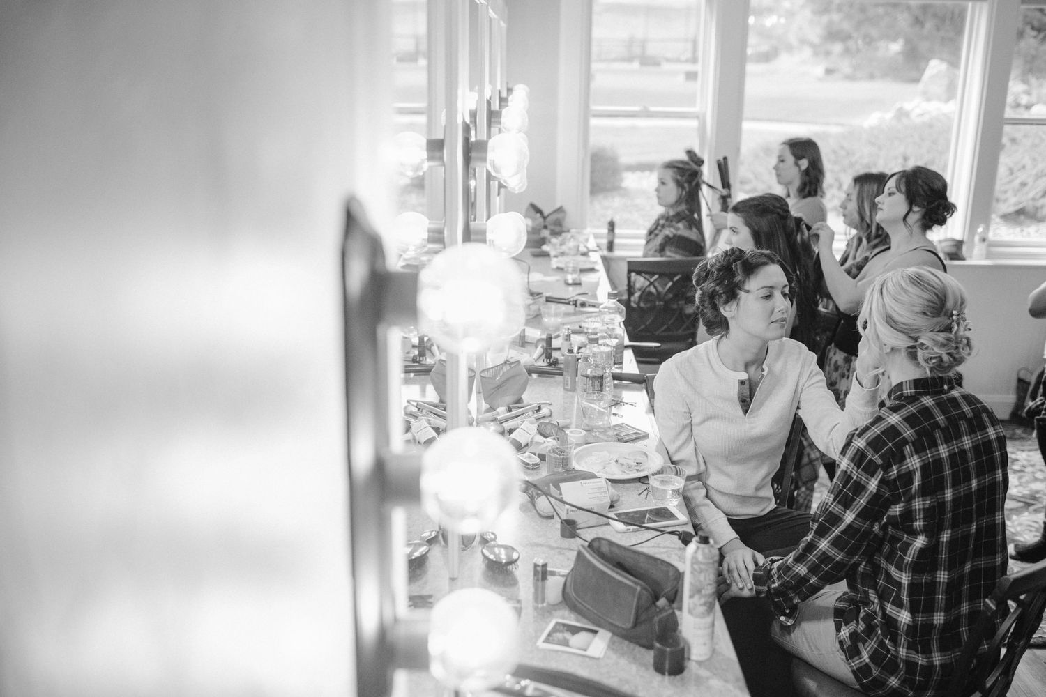 Black and white image of people getting ready at makeup stations with vanity lights.