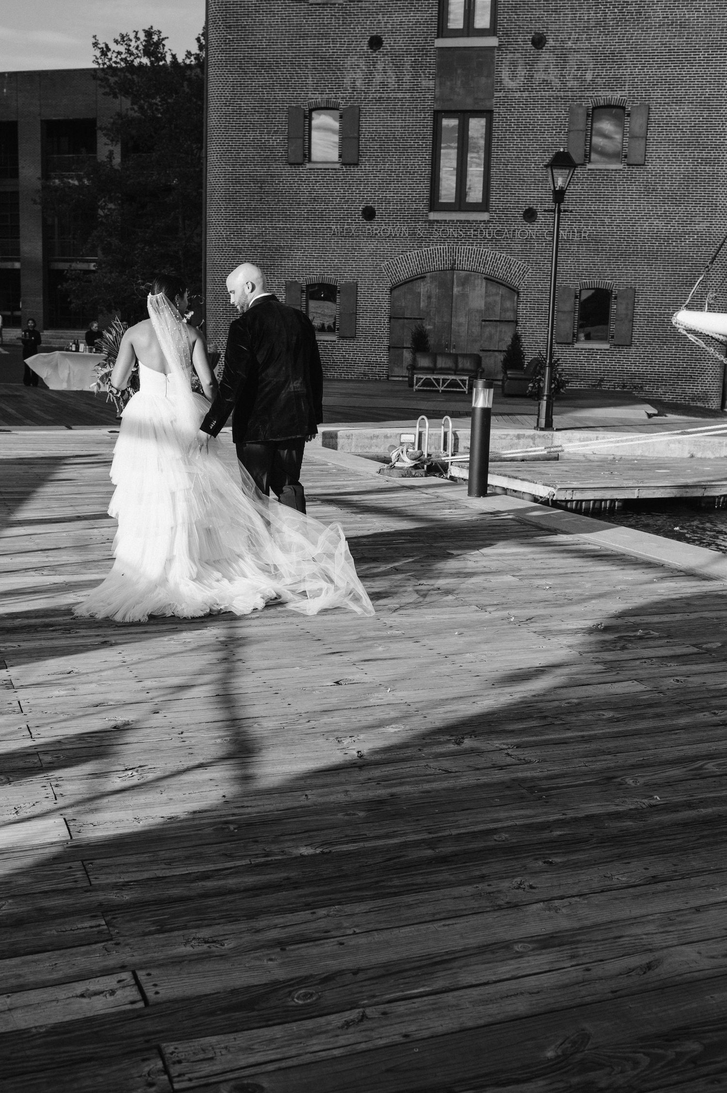 Black and white photo of a couple walking away on a wooden boardwalk near a historic brick building.