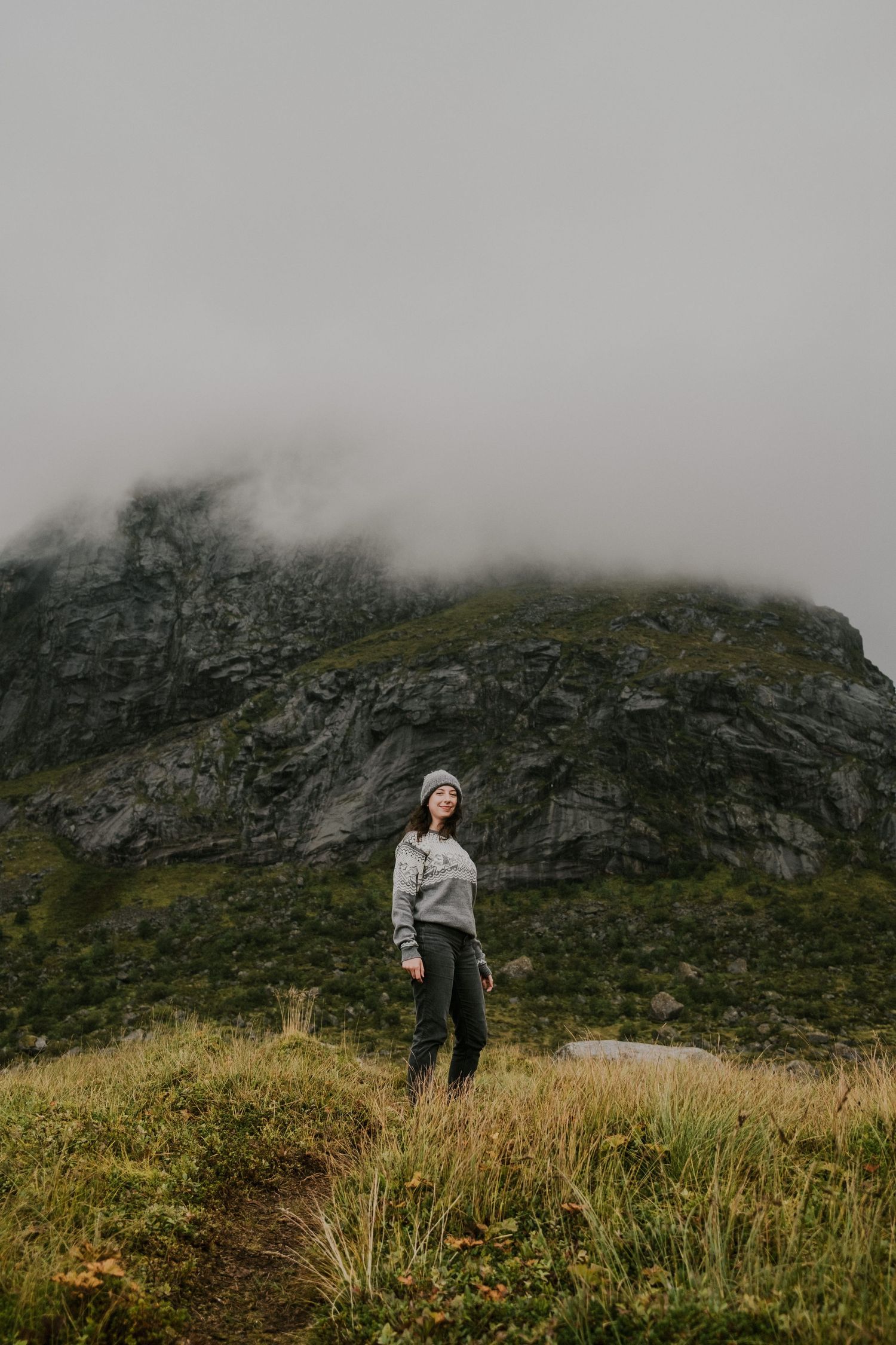 Winter hiker on grassy slope beneath foggy mountain peak.