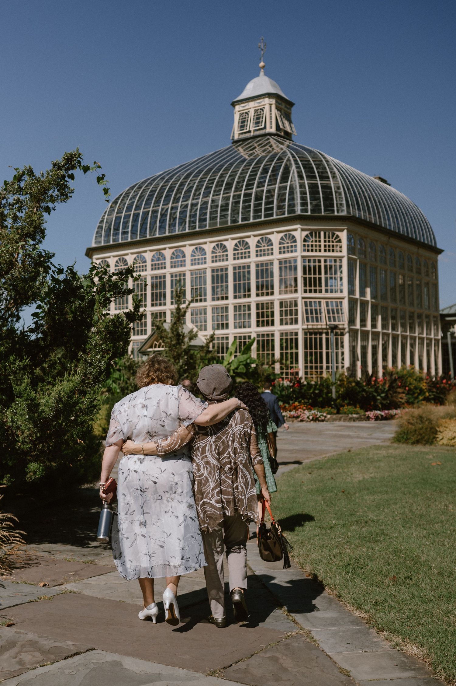 A couple walks together outside a Victorian-style greenhouse with a domed glass roof.