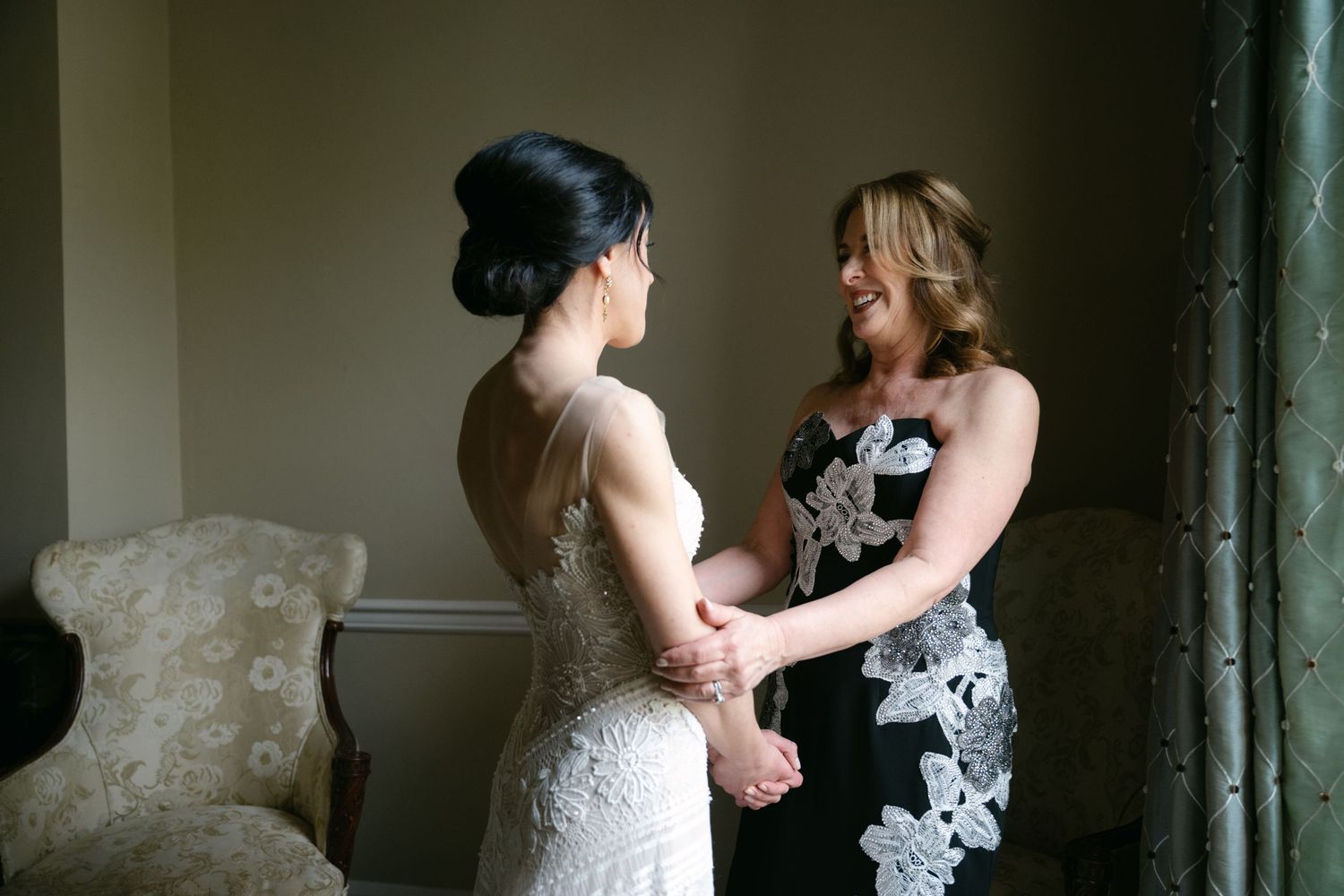 Two women sharing an emotional moment in formal dresses during a special occasion beside an elegant curtained window.