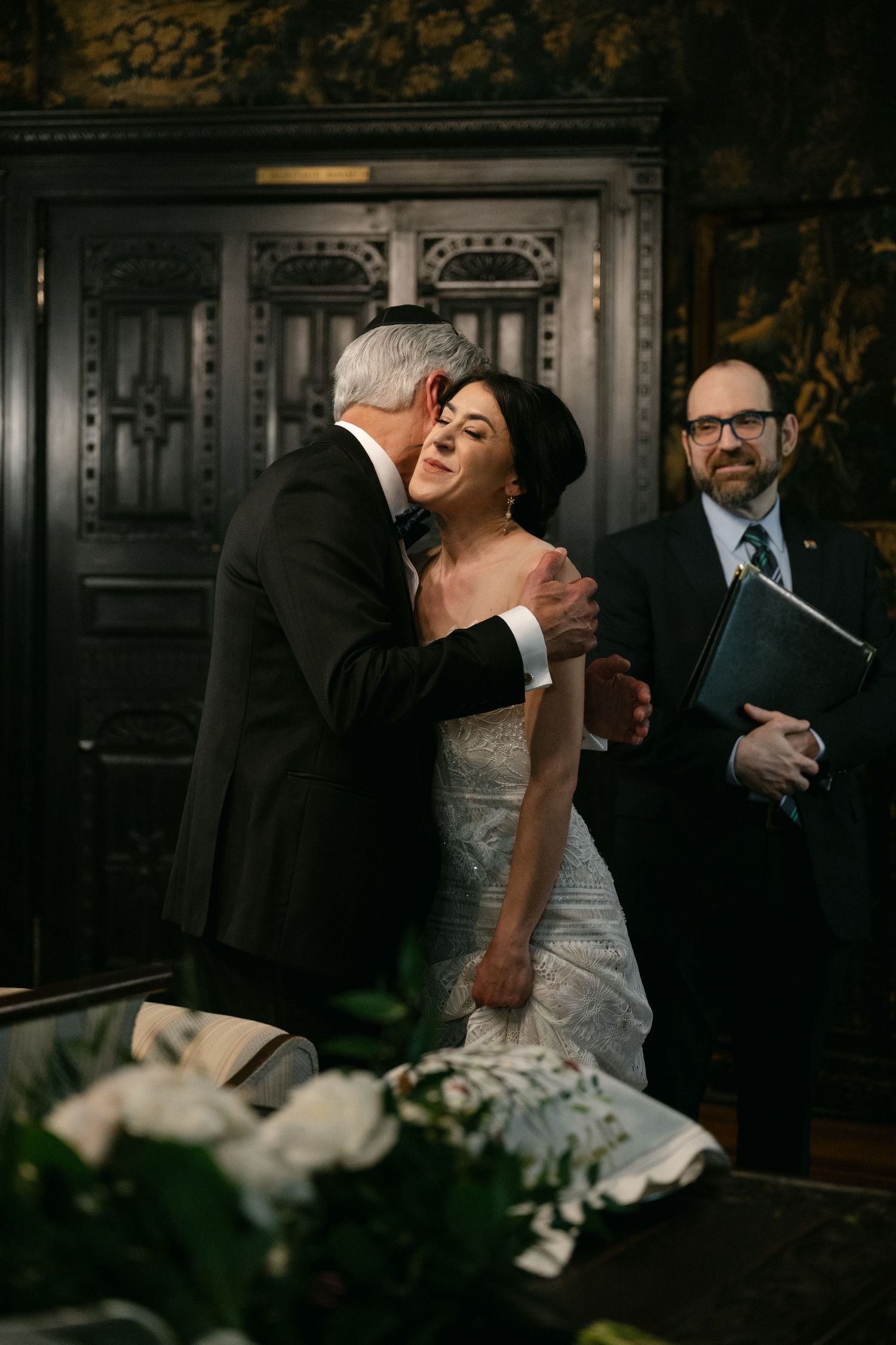 An emotional wedding ceremony moment captured in a dark wood-paneled room with ornate decor.