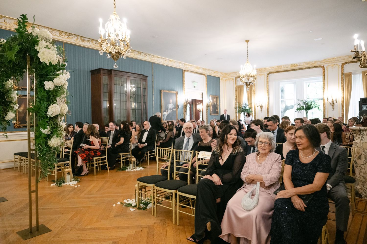 Wedding ceremony guests seated in rows of gold chairs in an elegant room with floral decorations.